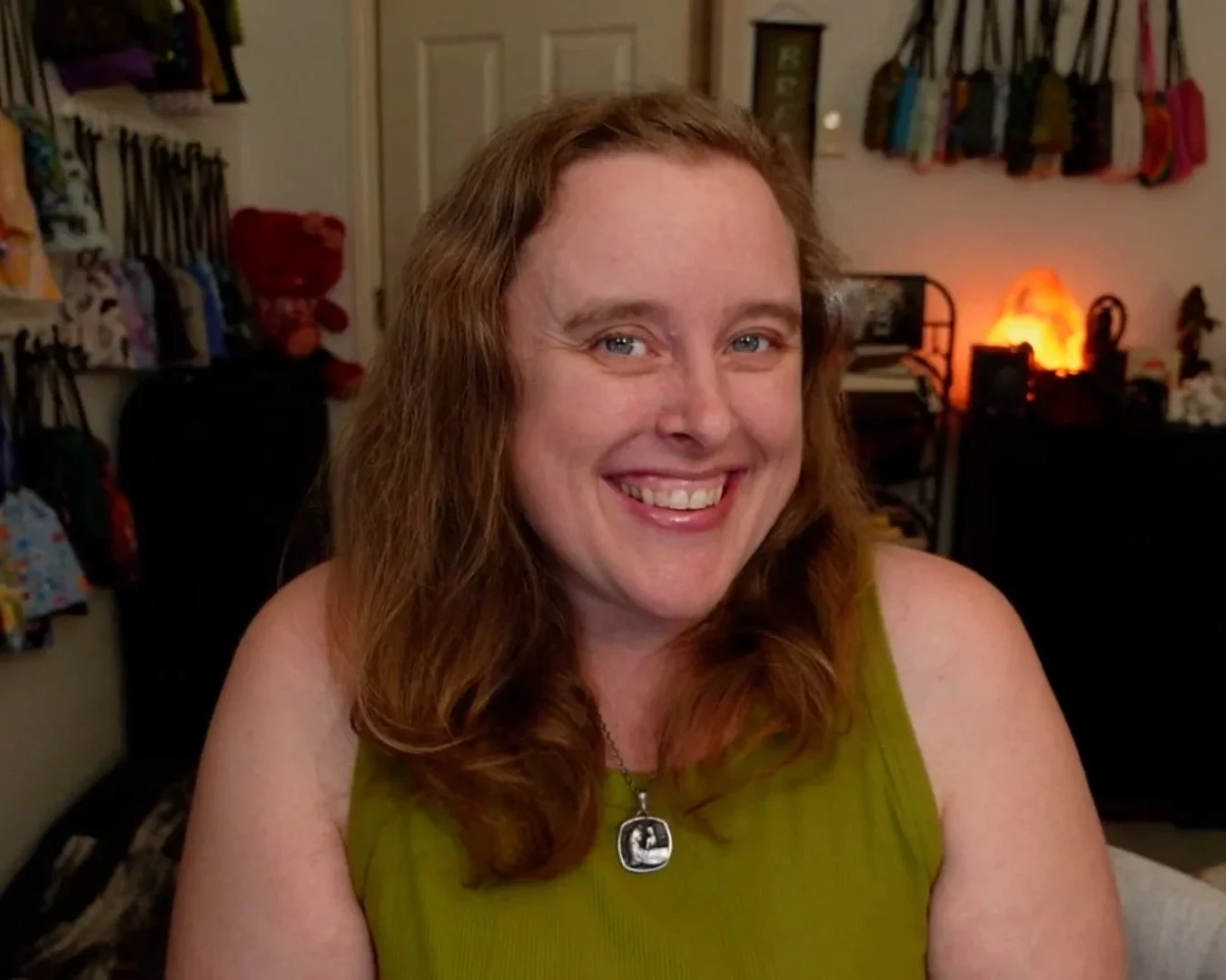 Lisa smiles at the viewer from her usual spot at her Tarot table. On the wall behind her, dozens of tarot and oracle decks can be seen hanging from the walls.