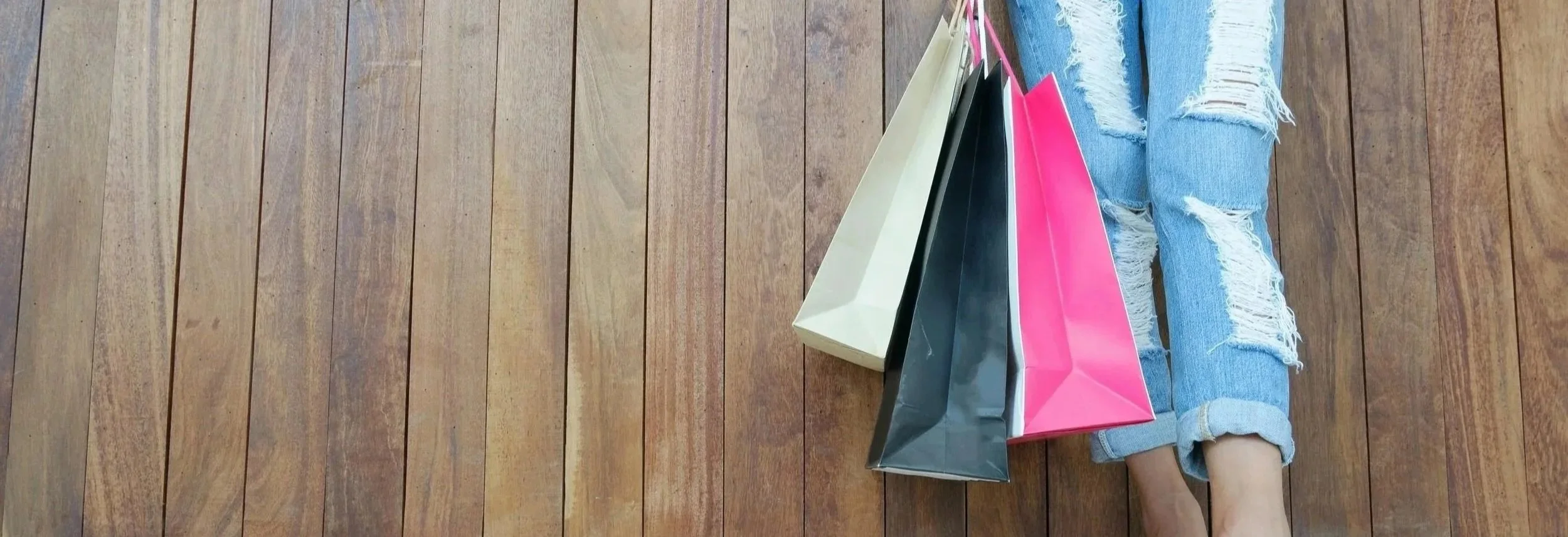 Person sitting on wooden floor with ripped jeans, holding pink, black, and cream shopping bags.