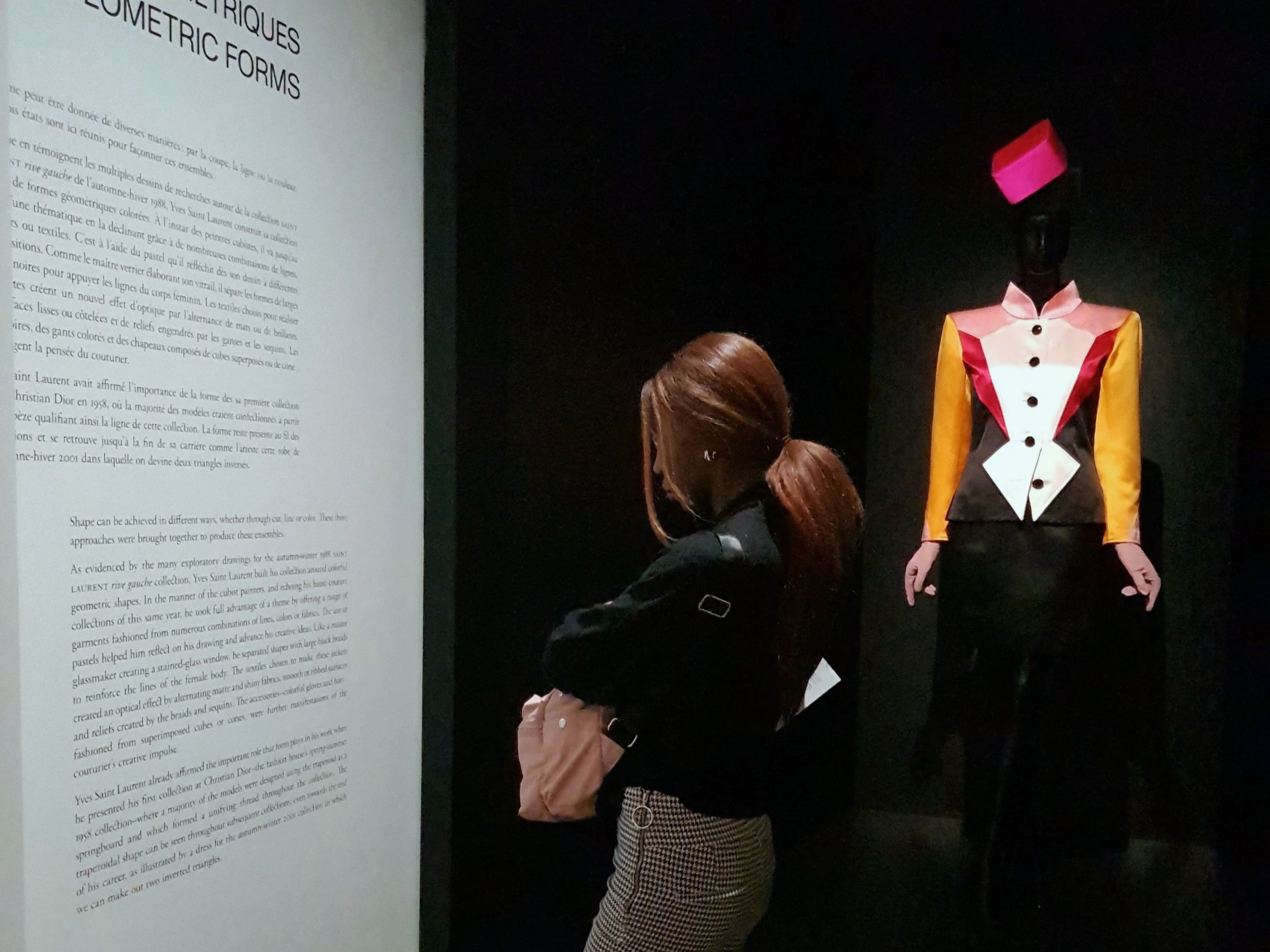 A woman with long brown hair, wearing a black and beige plaid outfit, stands in front of a display with a black mannequin dressed in a colorful geometric fashion. The display has a white informational panel on the left.