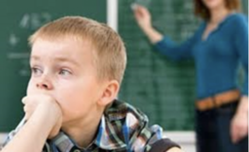 Niño distraído en clase mientras la profesora está al fondo junto a la pizarra.