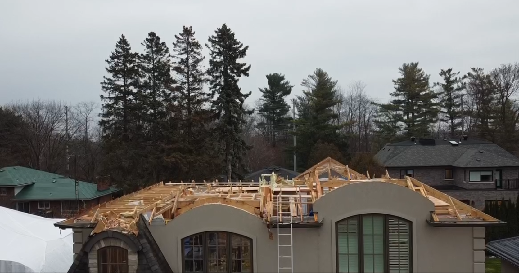 House under construction with a wooden roof framework, surrounded by trees and neighboring houses, on a cloudy day.
