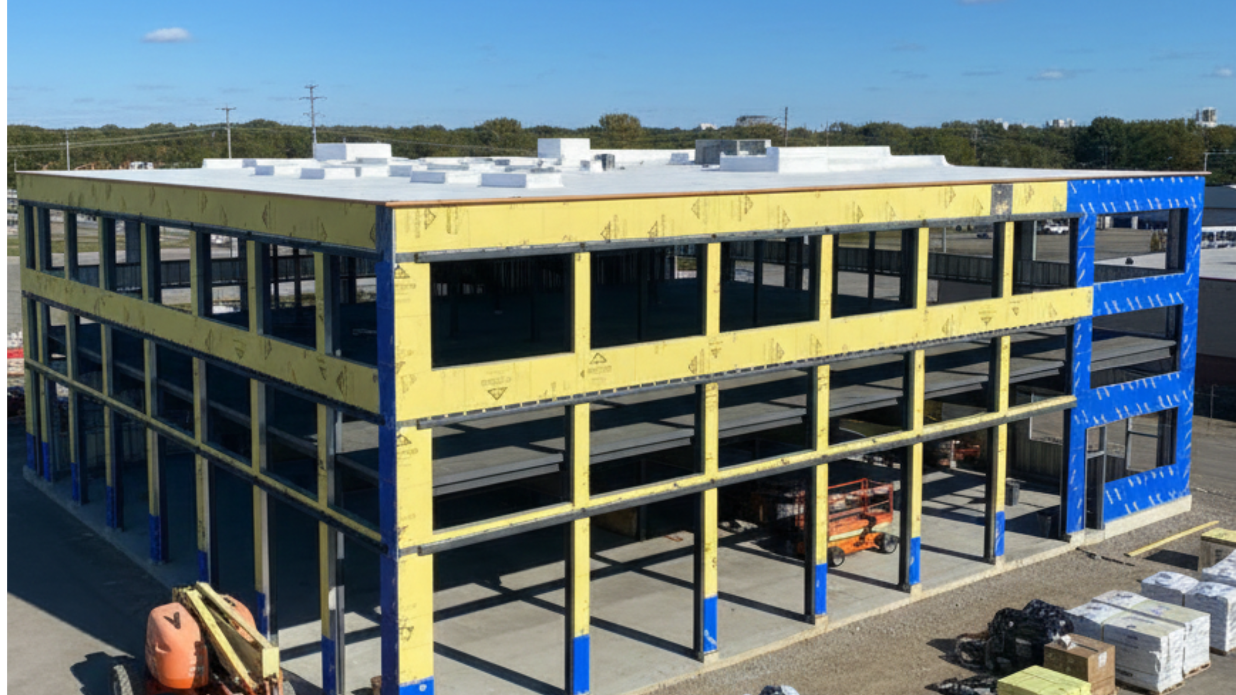 Construction site of a multi-story building with yellow and blue framing, partially built with open walls, situated on a concrete foundation with construction equipment and materials around.