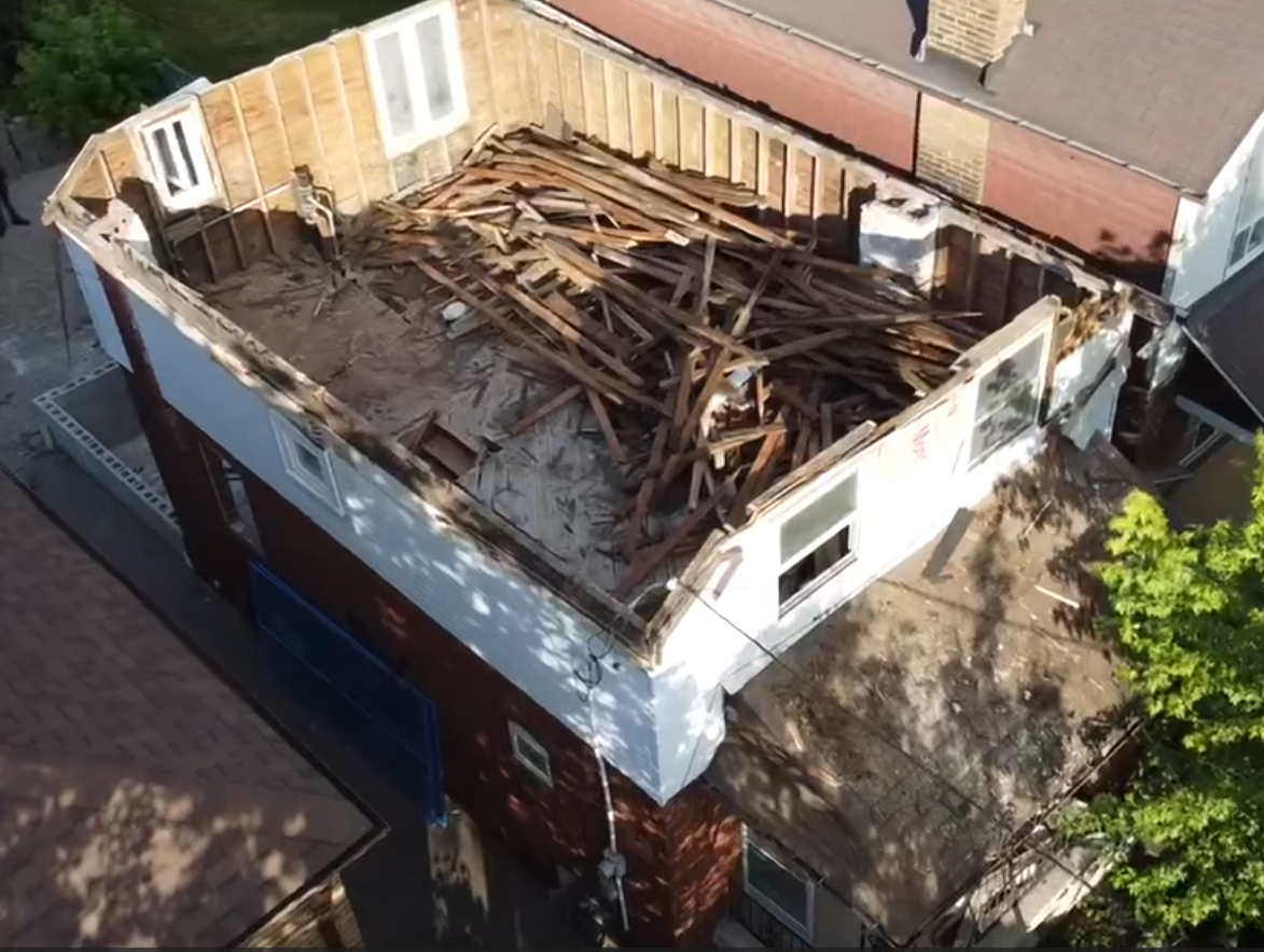Aerial view of a two-story building with its roof partially demolished, revealing wooden beams and debris inside.