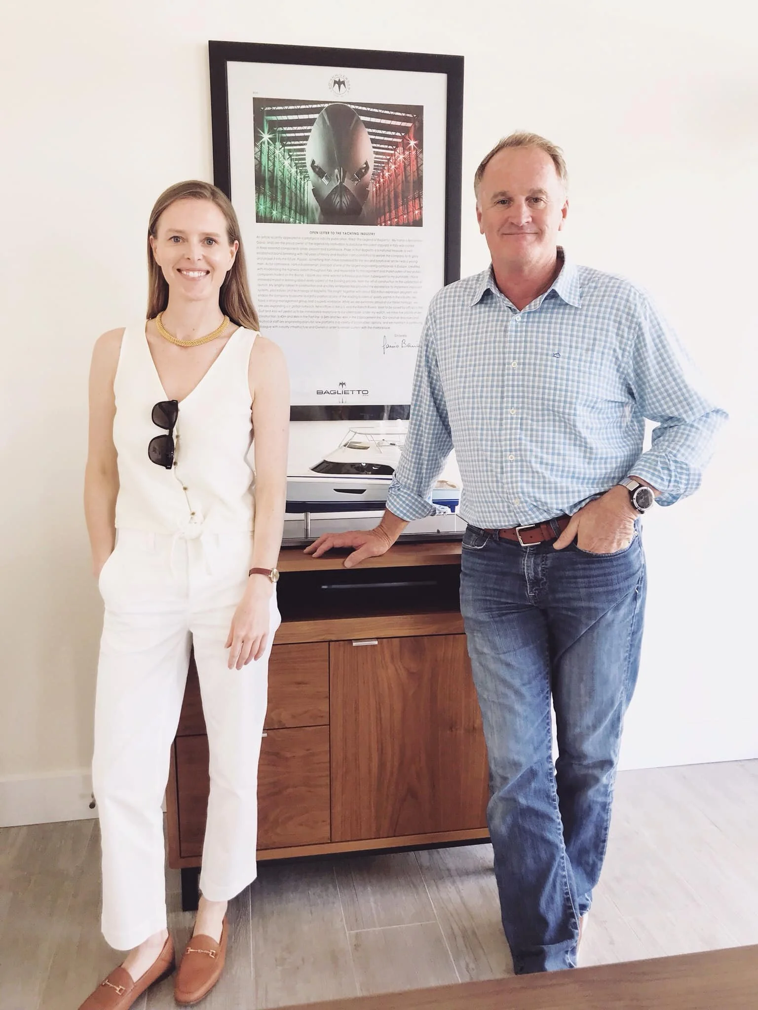 Two people standing in front of a framed artwork of a superhero helmet with a Bat logo. The woman on the left is wearing a white sleeveless jumpsuit, sunglasses hanging from her top, and brown shoes. The man on the right is dressed in a blue and white checkered shirt, blue jeans, and a watch, with his hand in his pocket.