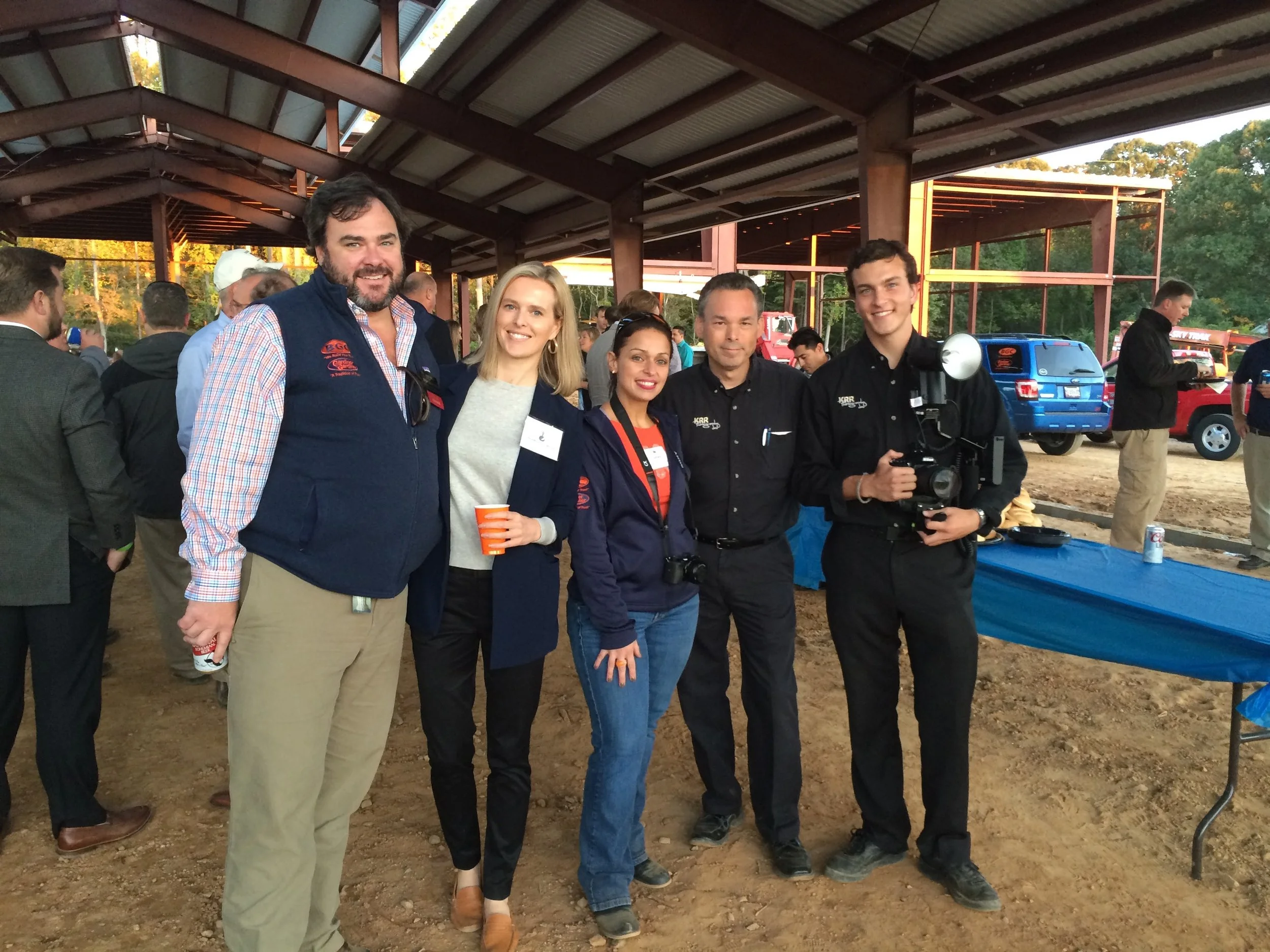 Group of five people standing together at an outdoor event, smiling for the photo. They are under a large open shelter with cars and other people in the background.
