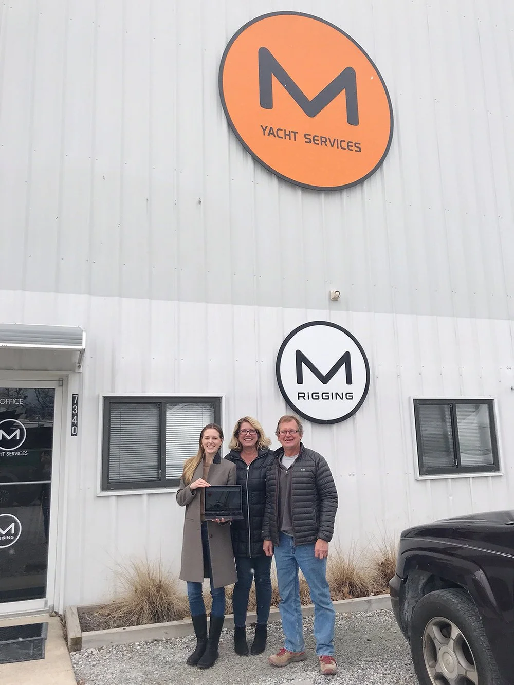 Three people standing outside a building with signs for Yacht Services and Rigging, smile for a photo, with a black vehicle partially visible in the foreground.