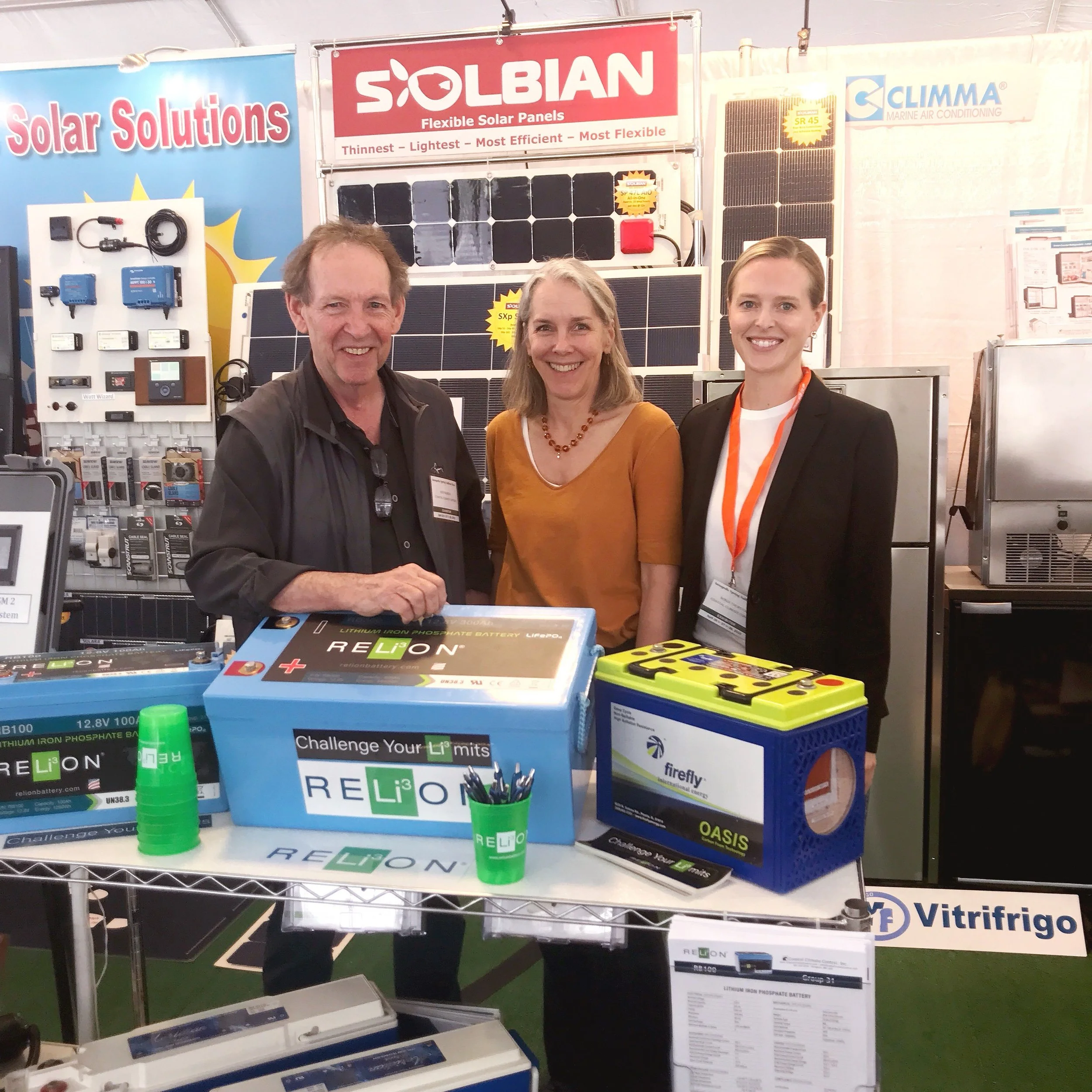 Three people standing behind a table with various battery and solar products at a trade show or exhibition. They are smiling and posing for the photo, with solar panels and promotional banners in the background.