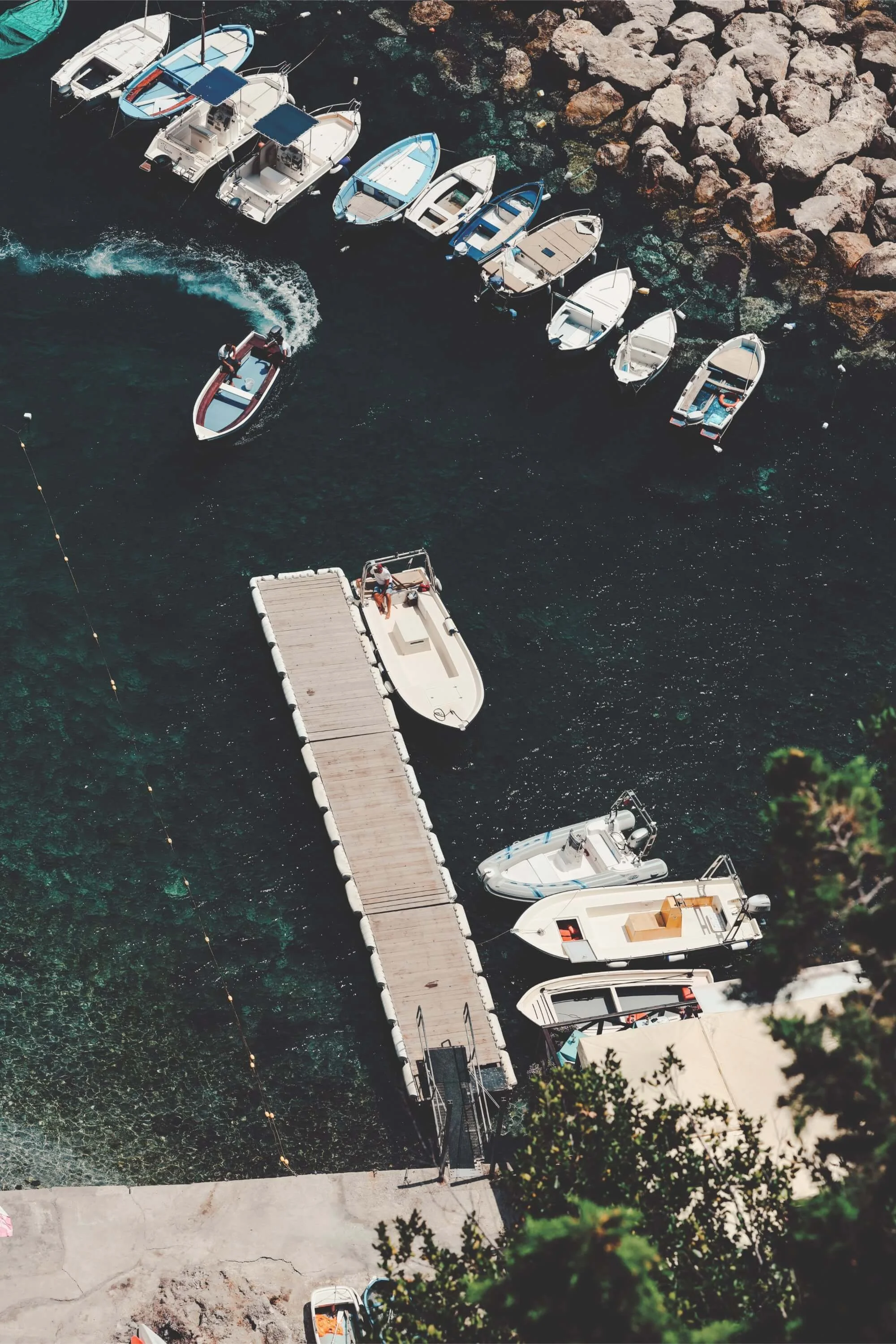 An aerial view of a marina featuring multiple boats docked along a pier extending into dark water. Some boats are anchored nearby, and a person is seen on a small boat navigating away from the dock. The shoreline is lined with large rocks and part of a paved walkway is visible at the bottom of the image.