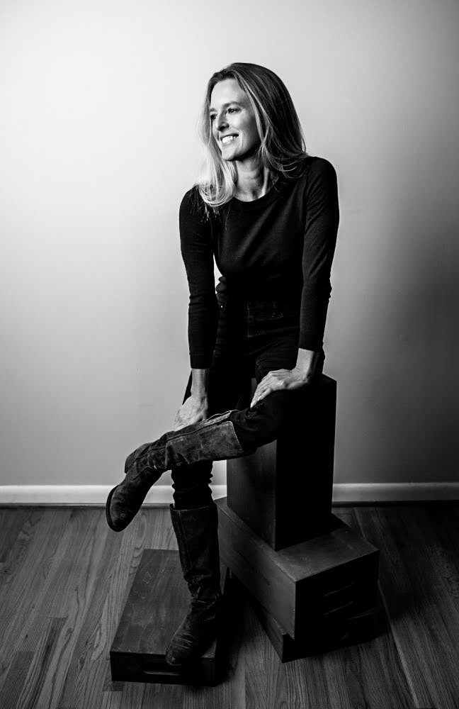 Black and white photo of a woman with shoulder-length hair smiling, sitting on stacked wooden blocks, wearing a long-sleeve top and knee-high boots, against a plain wall and hardwood floor background.