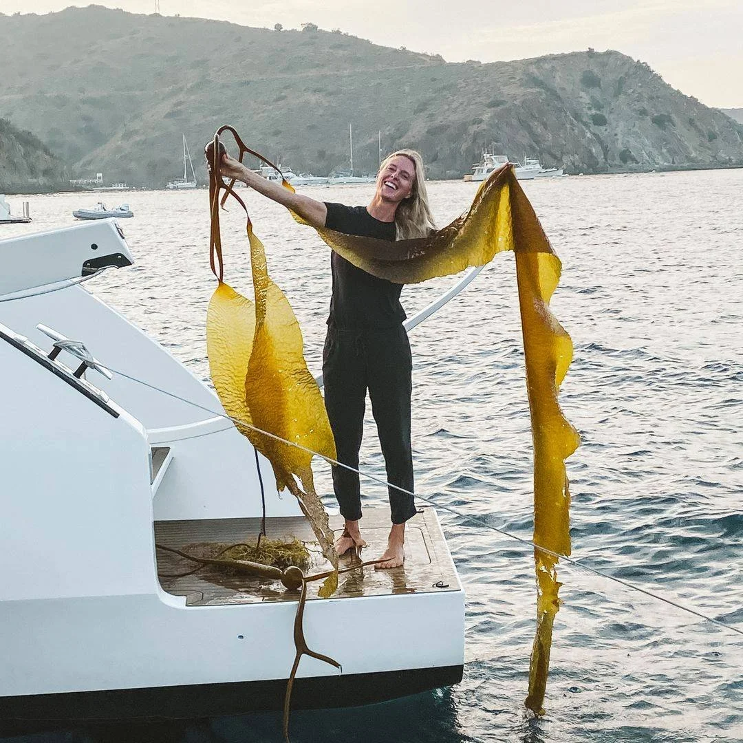 A woman smiling and holding a large piece of seaweed on a boat docked in a harbor with hilly islands and several boats in the background at sunset.