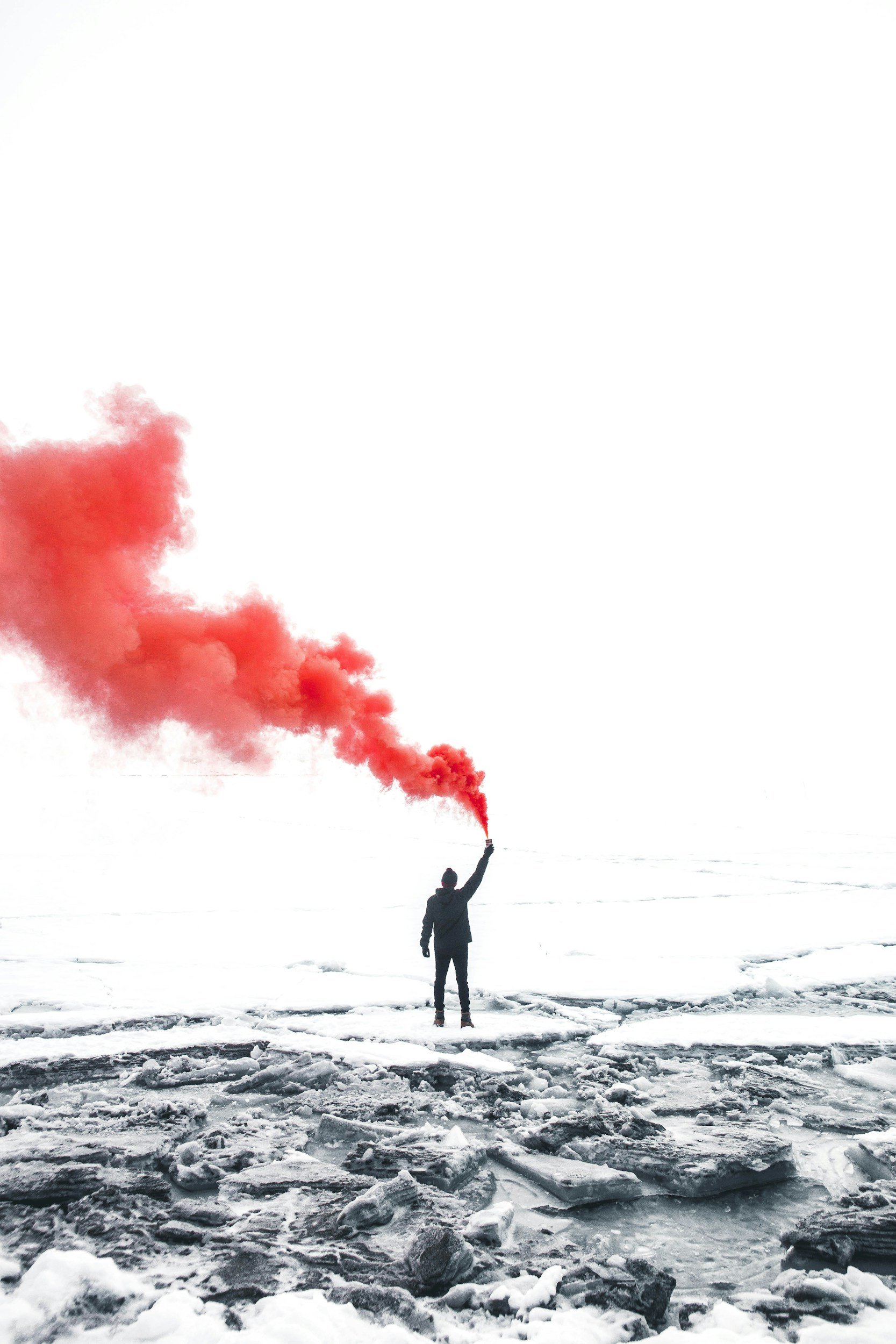 Person standing on icy shore holding red smoke flare, emitting a large cloud of red smoke in the air.