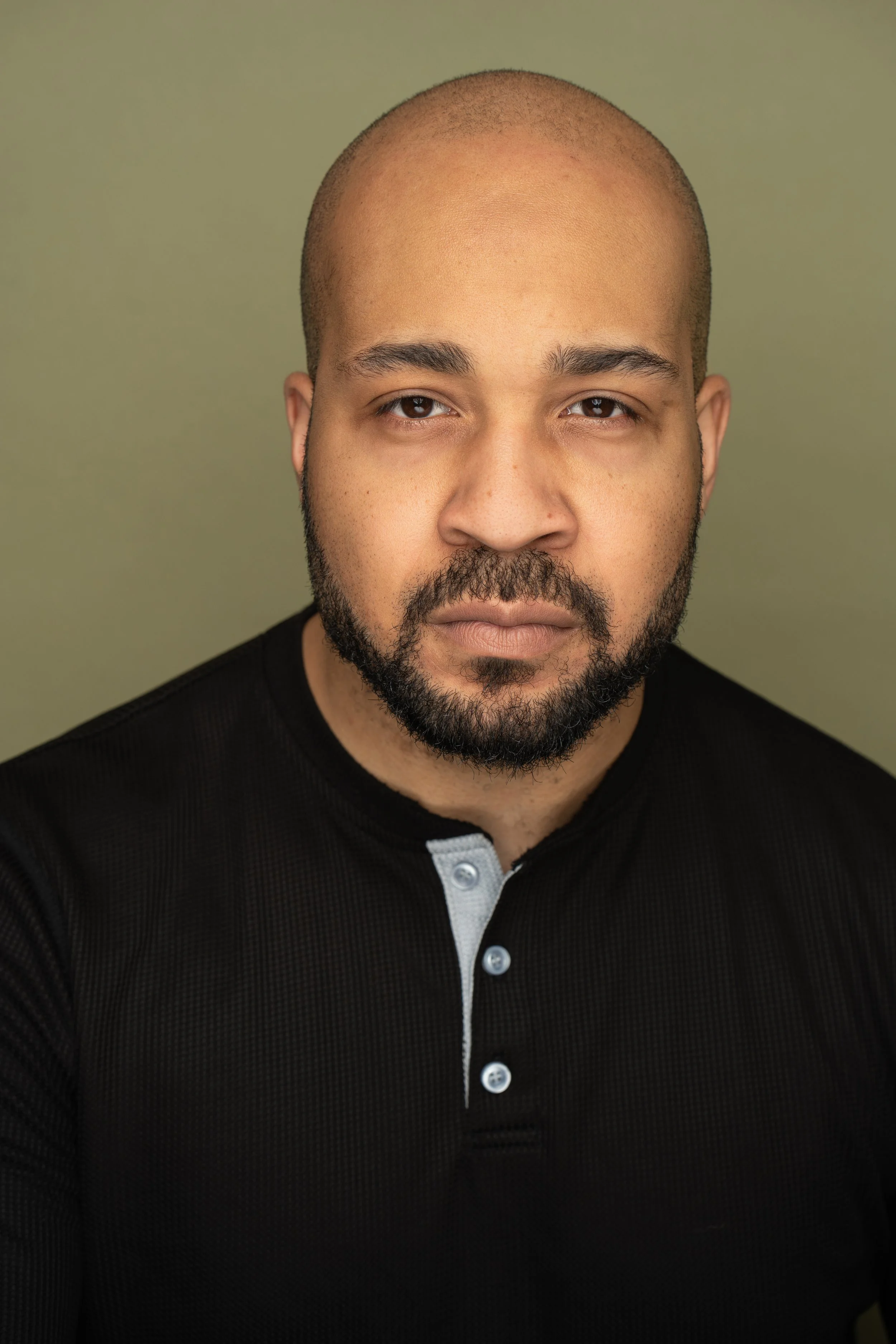 A man with a shaved head, beard, and mustache wearing a black shirt with a white button placket, looking directly at the camera against a plain light green background.