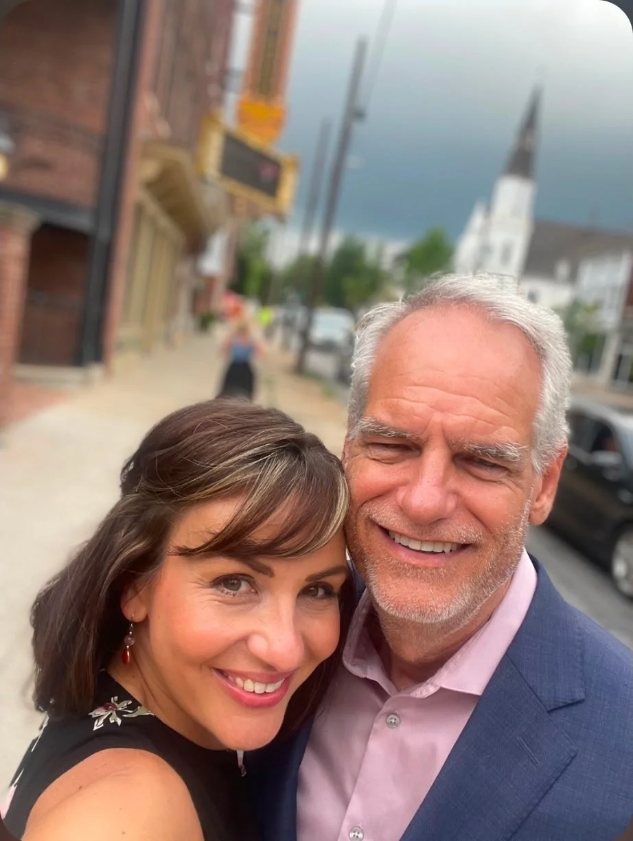 A smiling woman and man taking a selfie outdoors in a town with buildings, a church steeple, and a cloudy sky in the background.