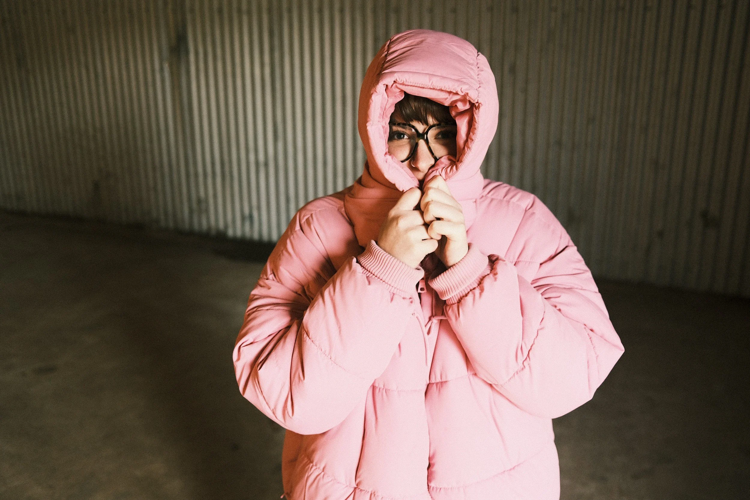 Woman in pink winter jacket with hood pulled over her head, standing in front of a corrugated metal wall, looking at the camera
