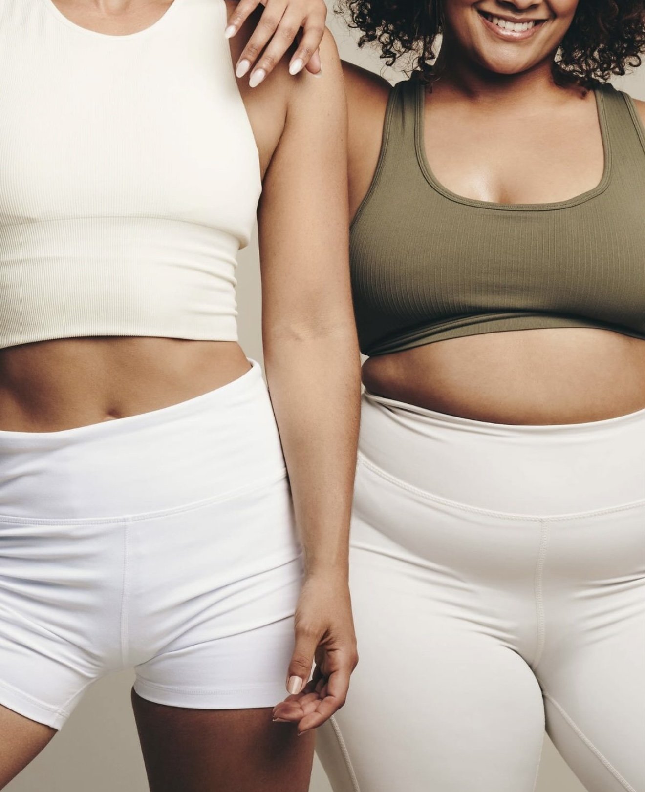 Two women standing together, one in a white crop top and shorts, the other in an olive green sports bra and white pants, smiling.