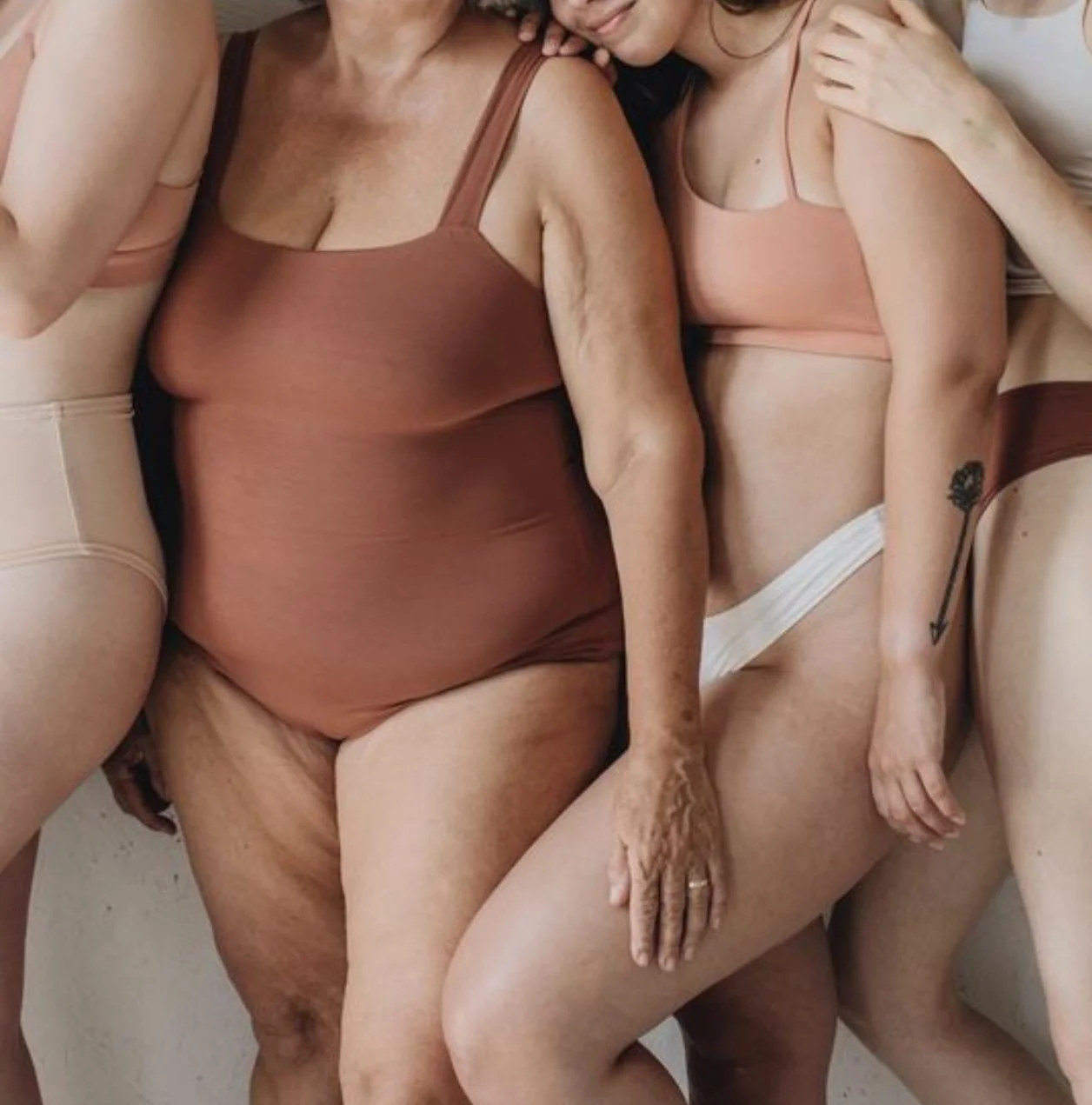 Close-up of four women in underwear, with one woman having a tattoo of an arrow on her arm, using a minimalist, natural style.