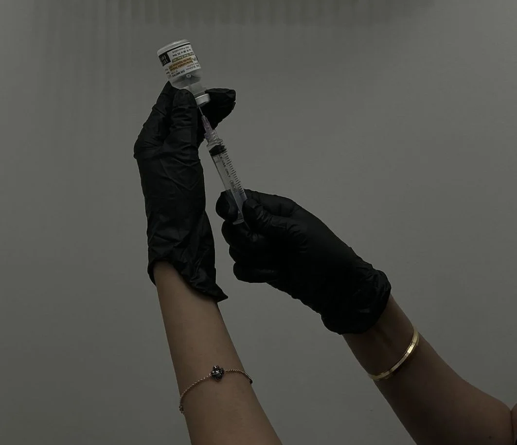 Hands wearing black gloves holding a vaccine vial and syringe against a plain gray background.