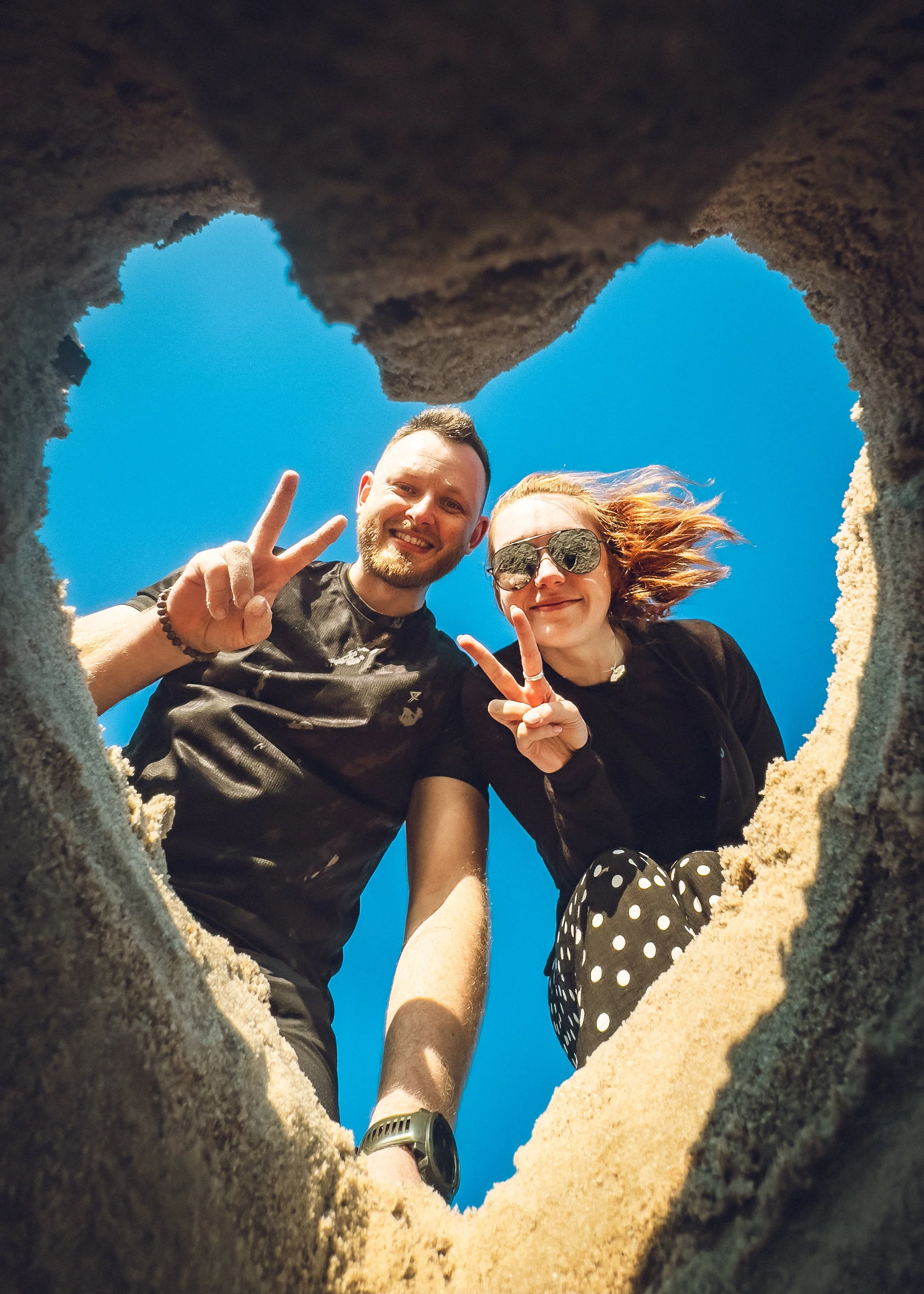 Two people taking a photo from inside a hole in the sand, looking down at the camera, with bright blue sky in the background. They are smiling and making peace signs.