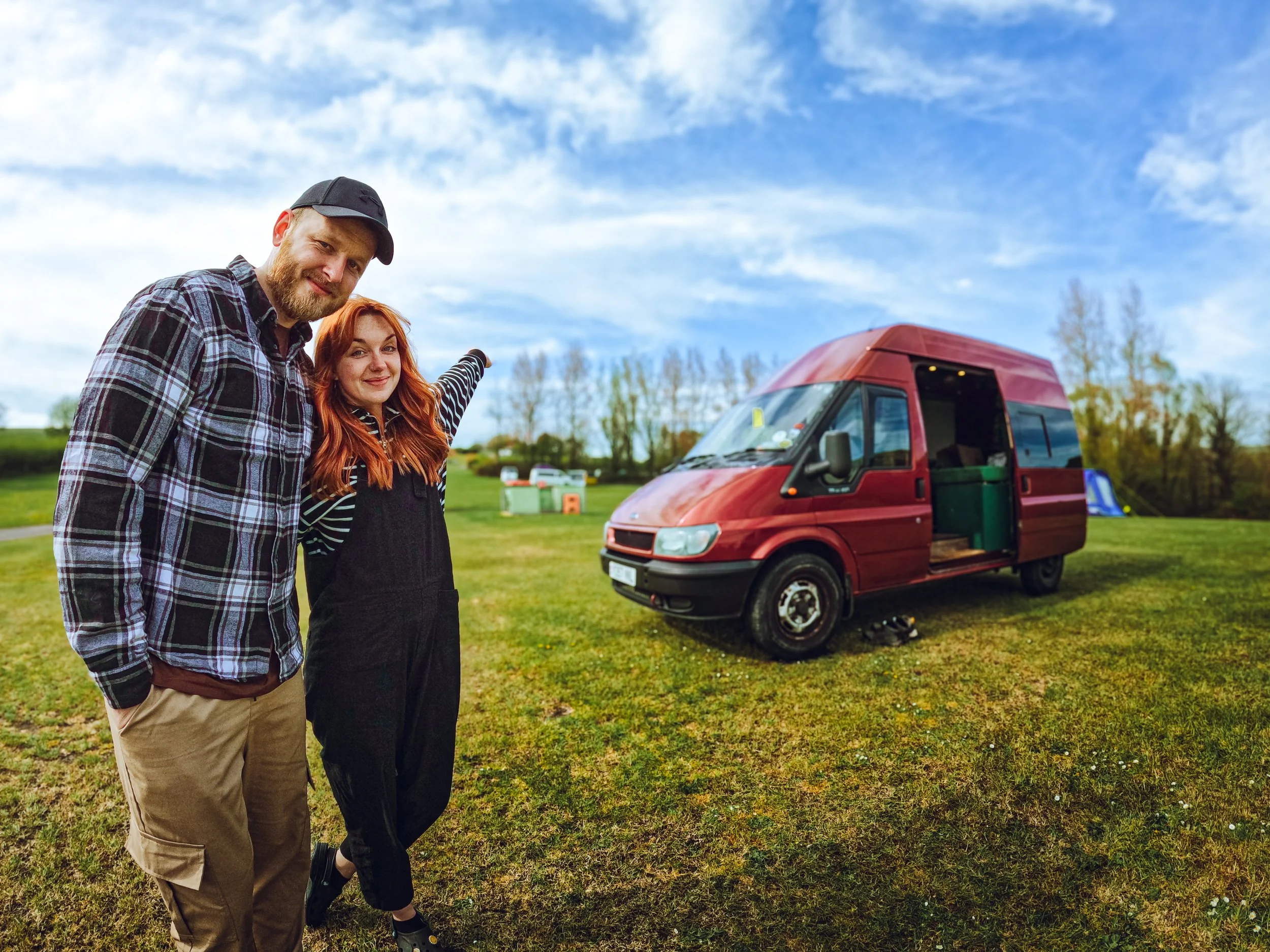 A man and woman standing on a grassy field near a red camper van, with the woman pointing at something in the distance.