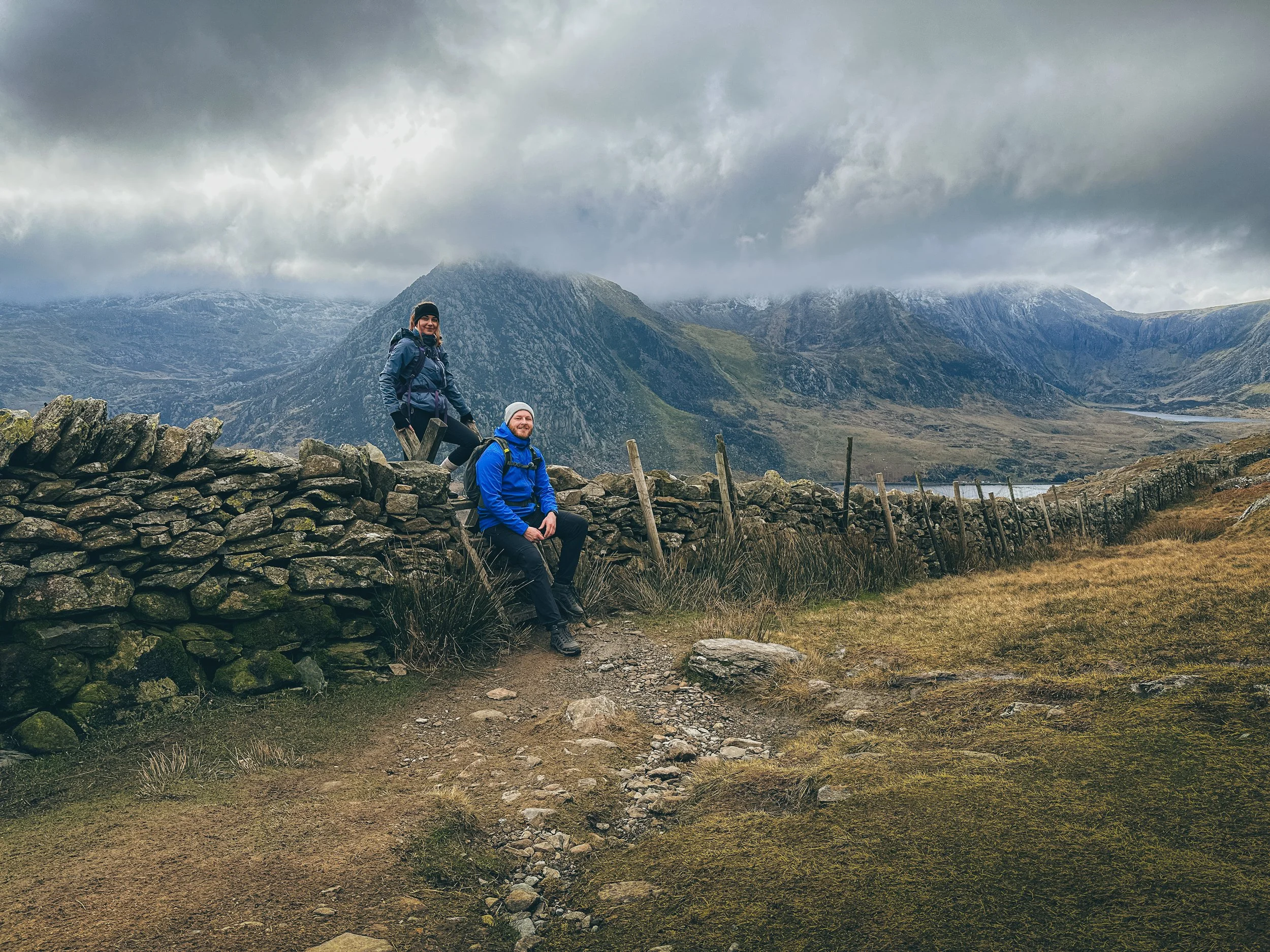 Two hikers in blue and black jackets sitting and standing on a stone wall in a mountainous landscape with cloudy sky and green valley.
