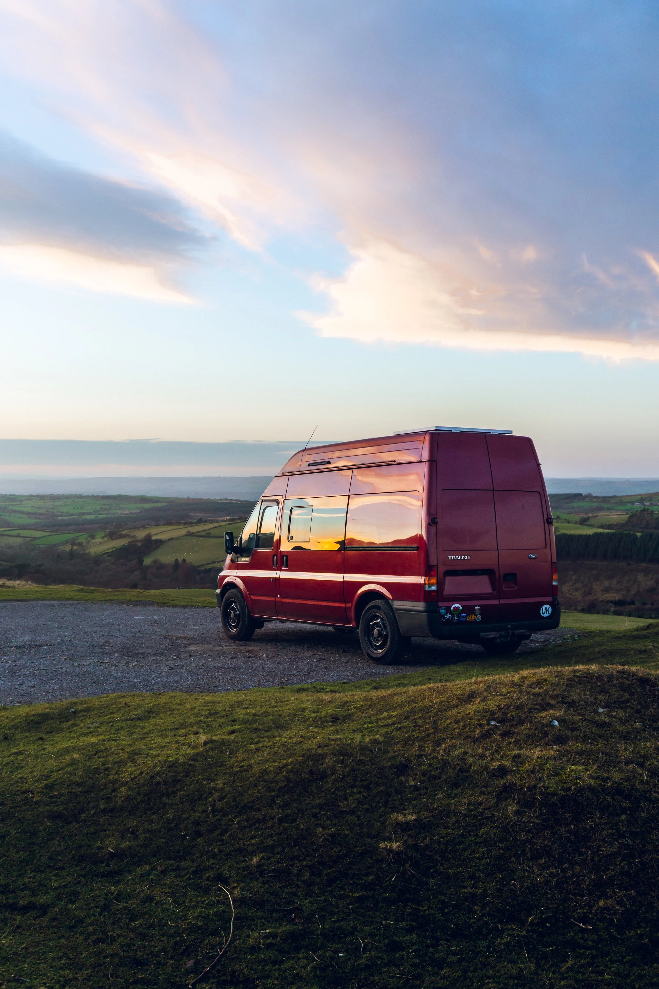 Red van parked on grass overlooking rolling hills at sunset with a colorful sky.