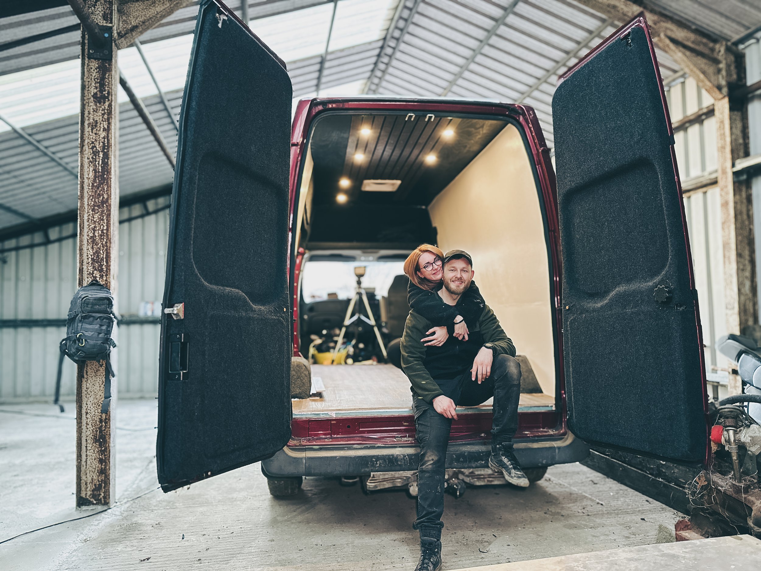A couple sitting at the back of an open cargo van inside a warehouse, with the woman hugging the man from behind.