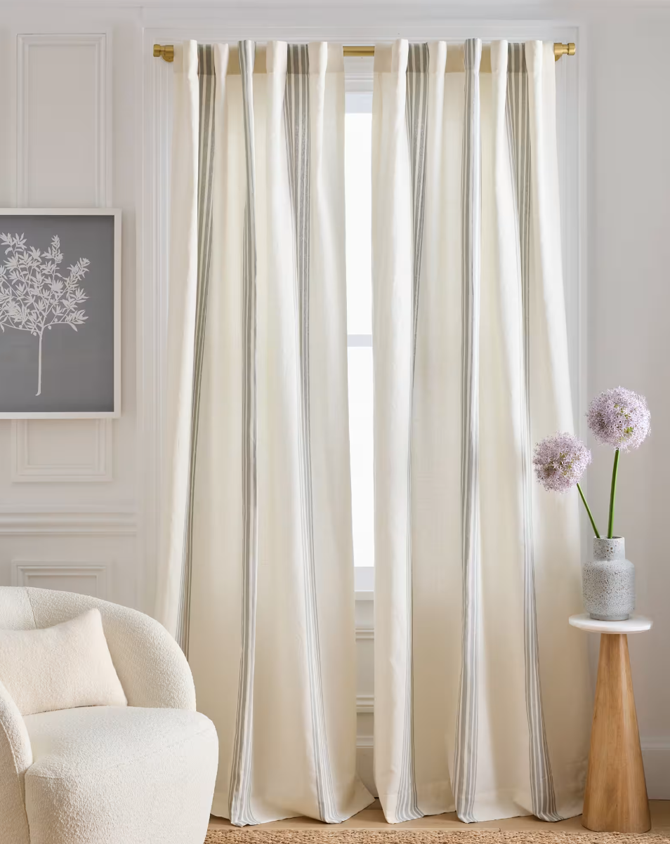 Light-colored curtains hanging in front of a window in a bright living room, with a cream armchair and a small side table with purple flowers in a vase.