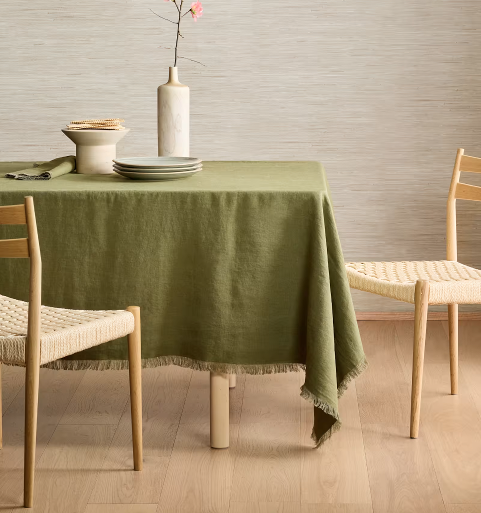 Dining table with a green tablecloth, wooden chairs with woven seats, a tall white vase with pink flowers, and a stack of plates on the table.