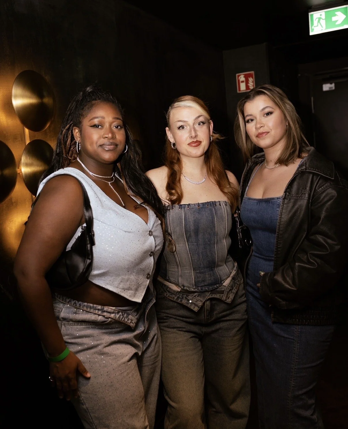 Three women posing together indoors at a social event, with dark walls, gold decorative elements, and visible emergency exit and fire safety signs behind them.