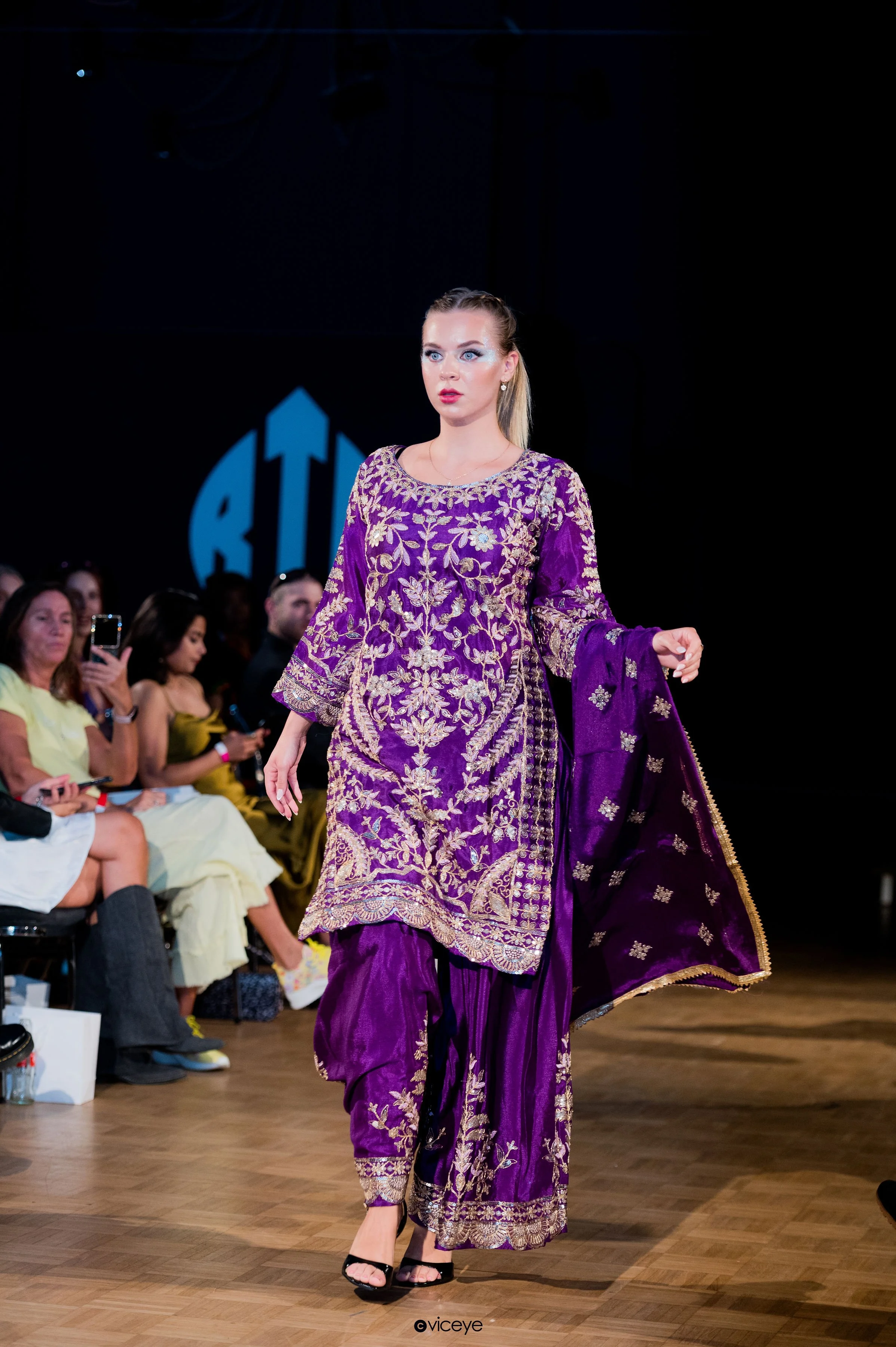 A female model walking down a runway in a purple and gold embroidered traditional outfit during a fashion show with seated audience on both sides.