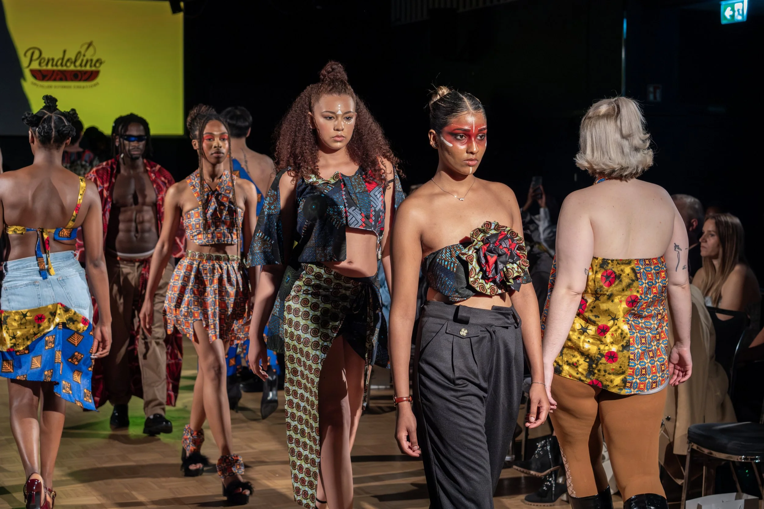 Models walk on runway during a fashion show, wearing colorful clothing with African-inspired patterns and styles, while audience members watch from the sides.