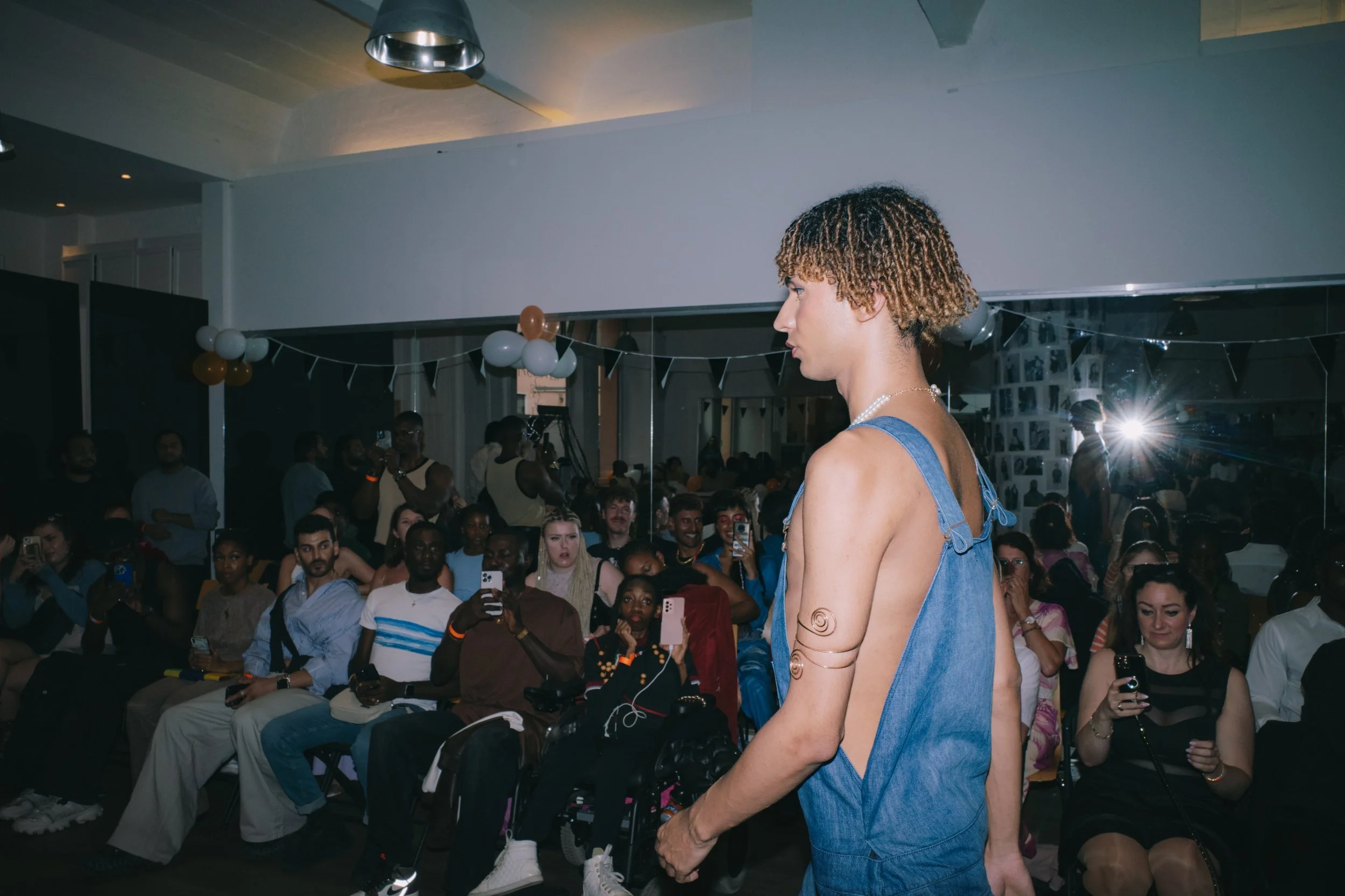 A woman in denim overalls walking in front of an audience at a fashion show, with people taking photos and videos of her.