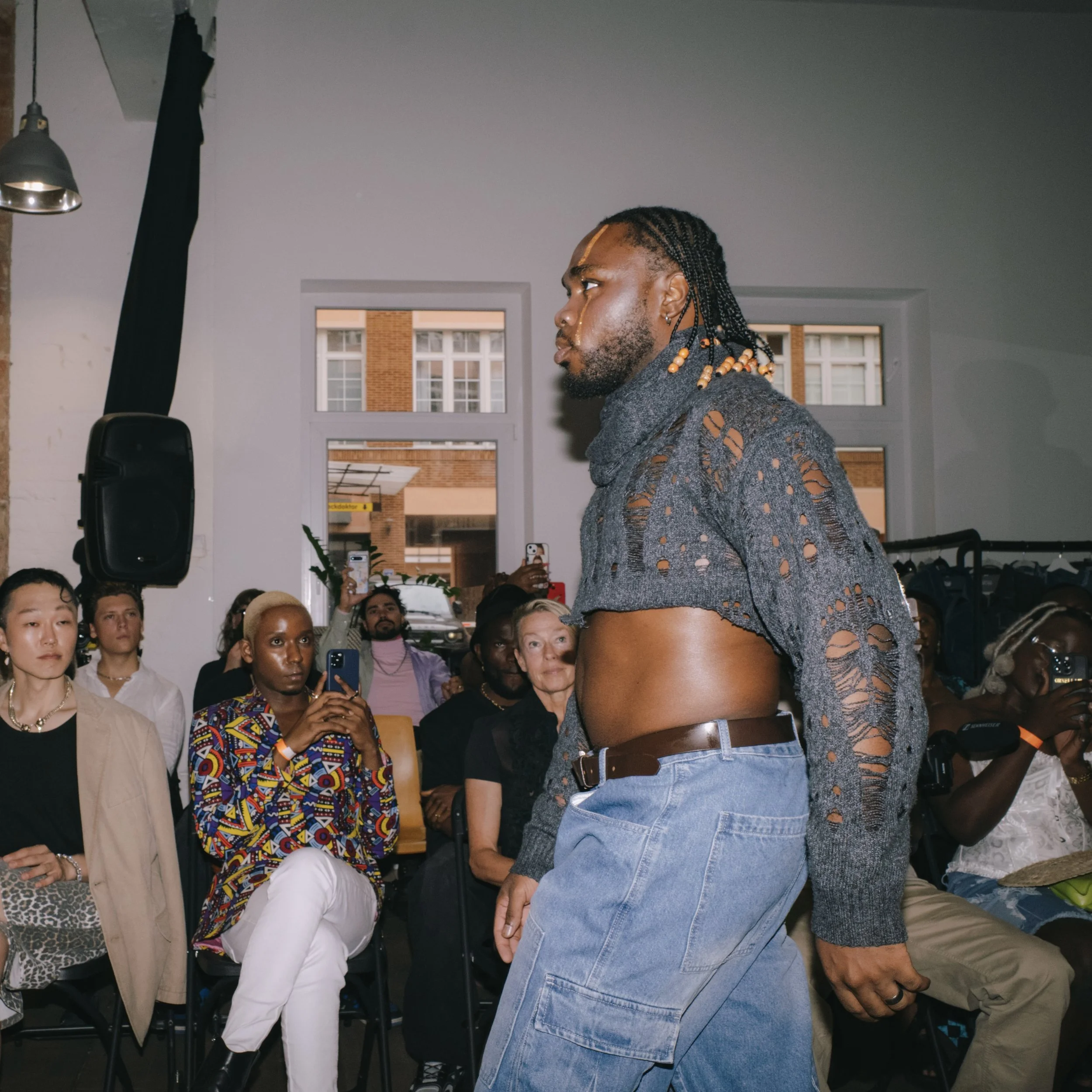 A male model walking on a runway wearing distressed gray sweater and loose jeans, with an audience watching.