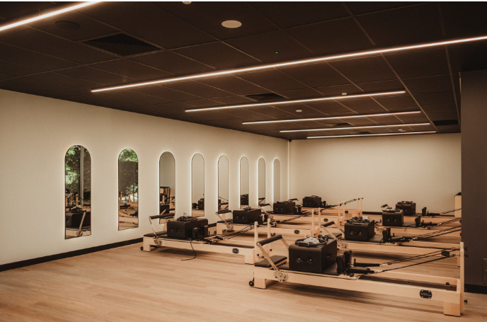 A spacious room with wooden flooring, featuring multiple reformer Pilates machines aligned in rows, illuminated by strip lights on a black ceiling, with a row of tall, arched mirrors along one wall reflecting the equipment and windows outside.