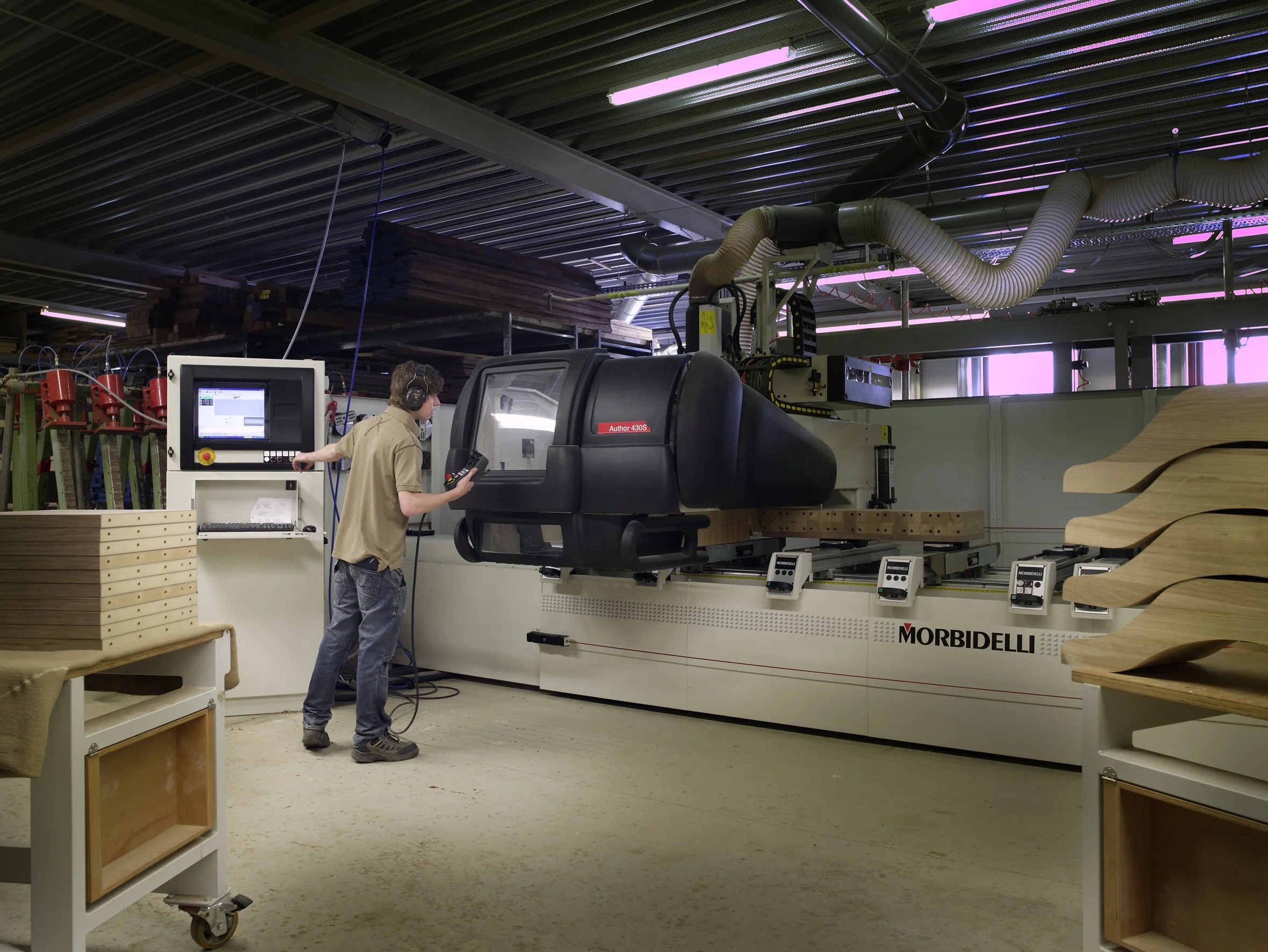 A worker in a wood workshop operates a CNC machine to shape wood panels. The machine is labeled Morbidei, and the worker wears headphones and is using a control panel.