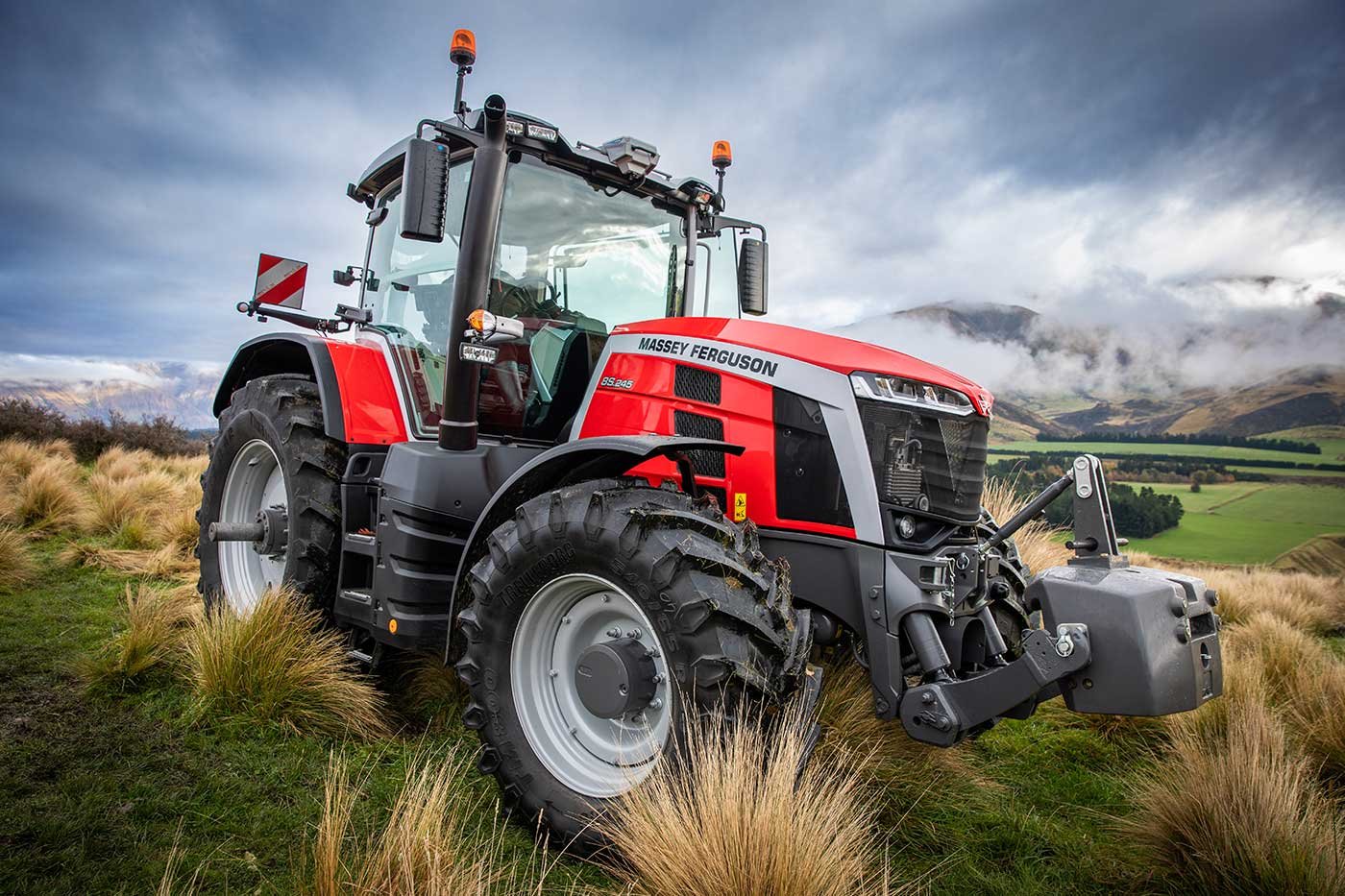 Red Massey Ferguson tractor on grassy field with hills and cloudy sky in background.