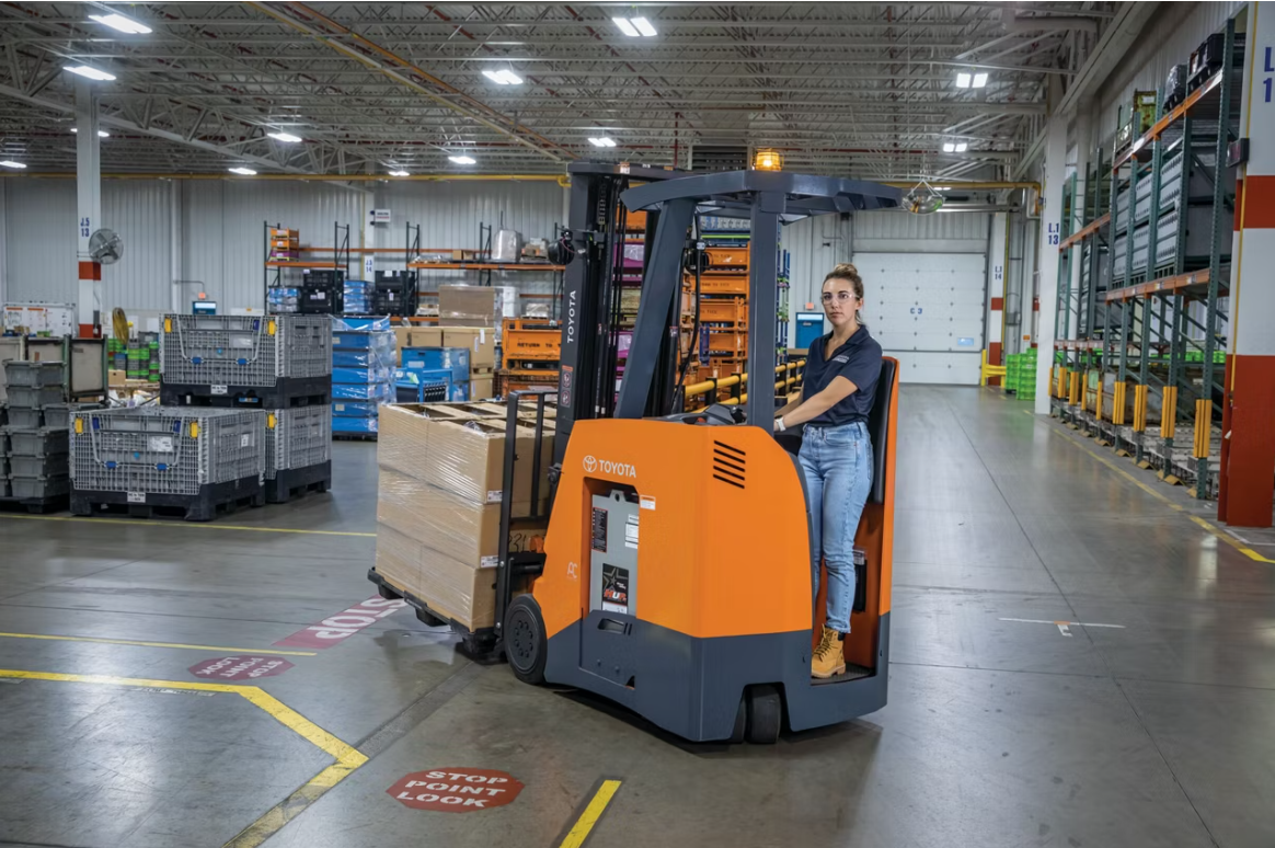A woman driving a Toyota electric pallet jack carrying a pallet of boxes inside a warehouse with shelves, storage bins, and industrial equipment.