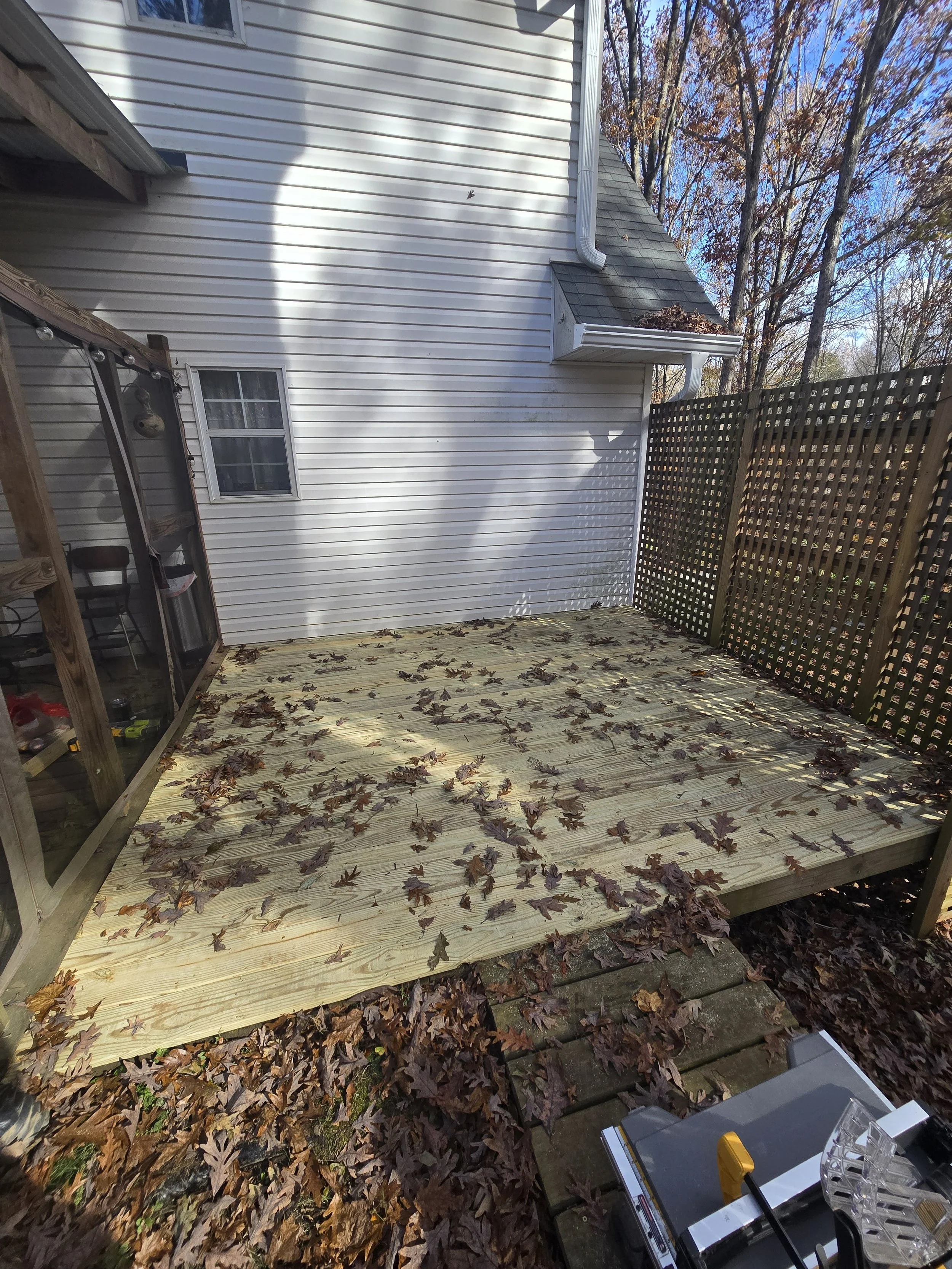 Newly built wooden deck attached to a house, covered with fallen autumn leaves, with a fenced yard and a small enclosed porch visible on the left.