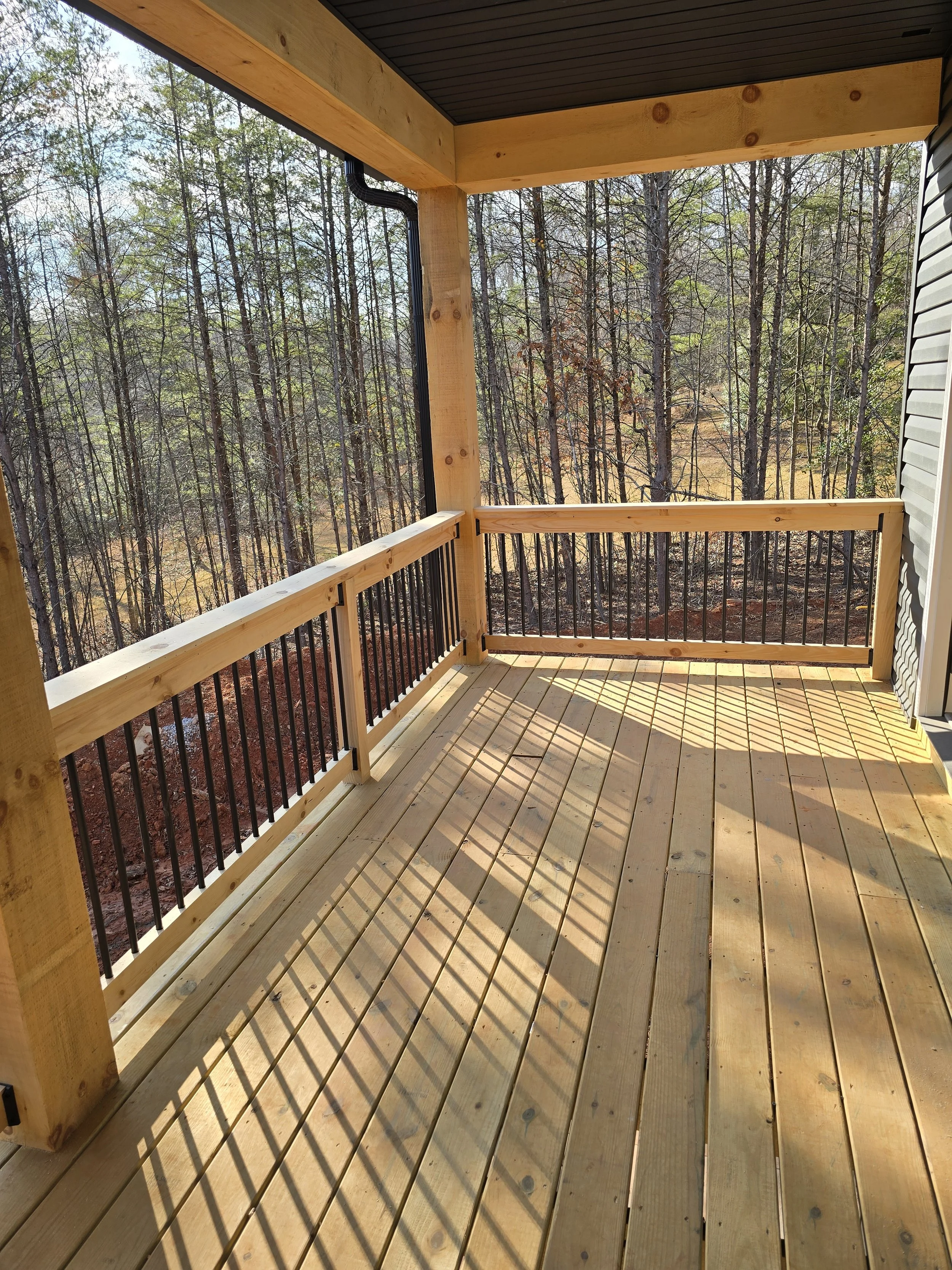 View of a newly built wooden balcony with black metal railings, overlooking a forested area with trees and a clear sky.