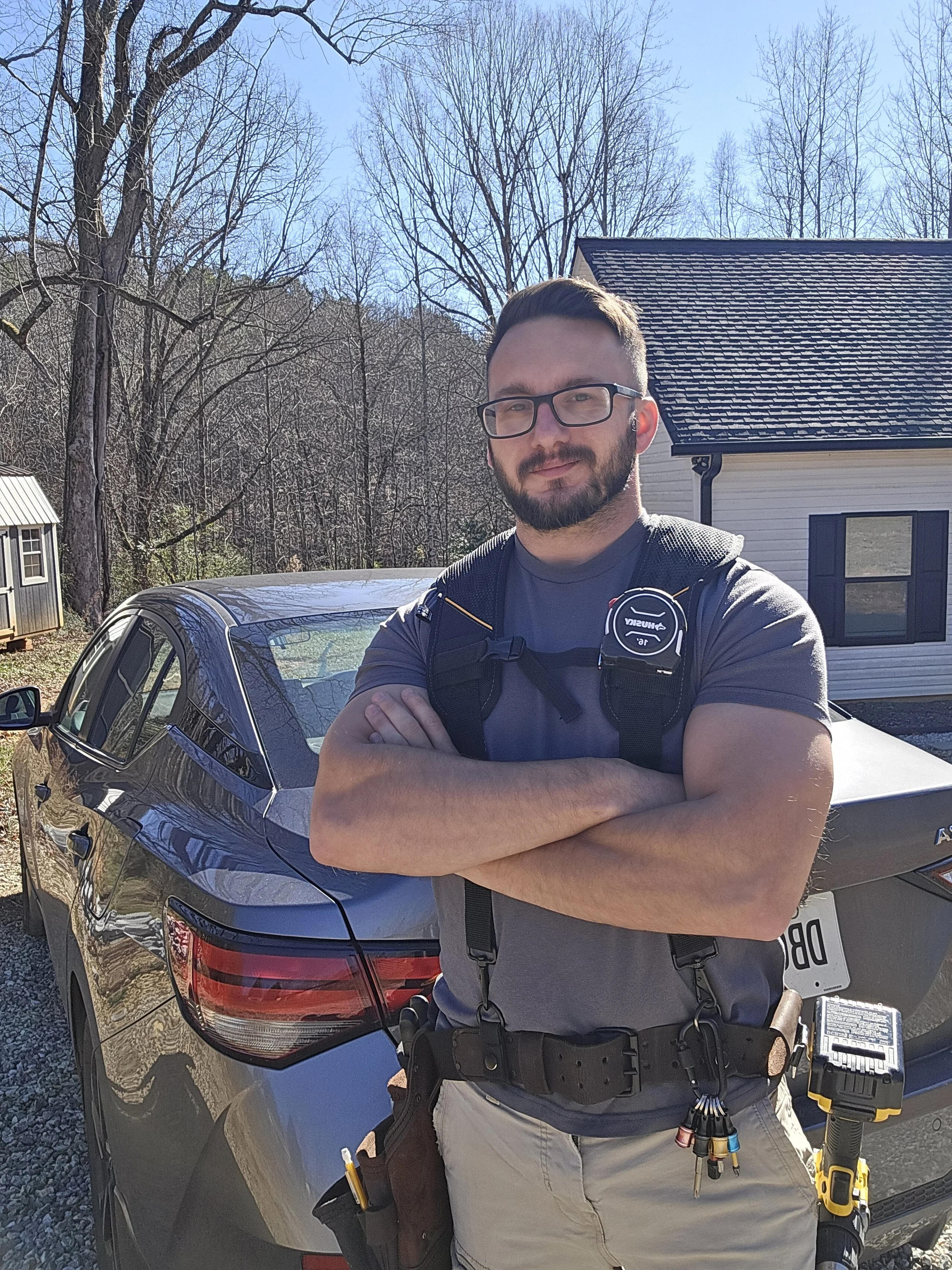 A man with glasses and a beard standing outdoors in front of a car, with arms crossed, wearing a tool belt and a harness, in a residential neighborhood with trees, houses, and a bright blue sky.