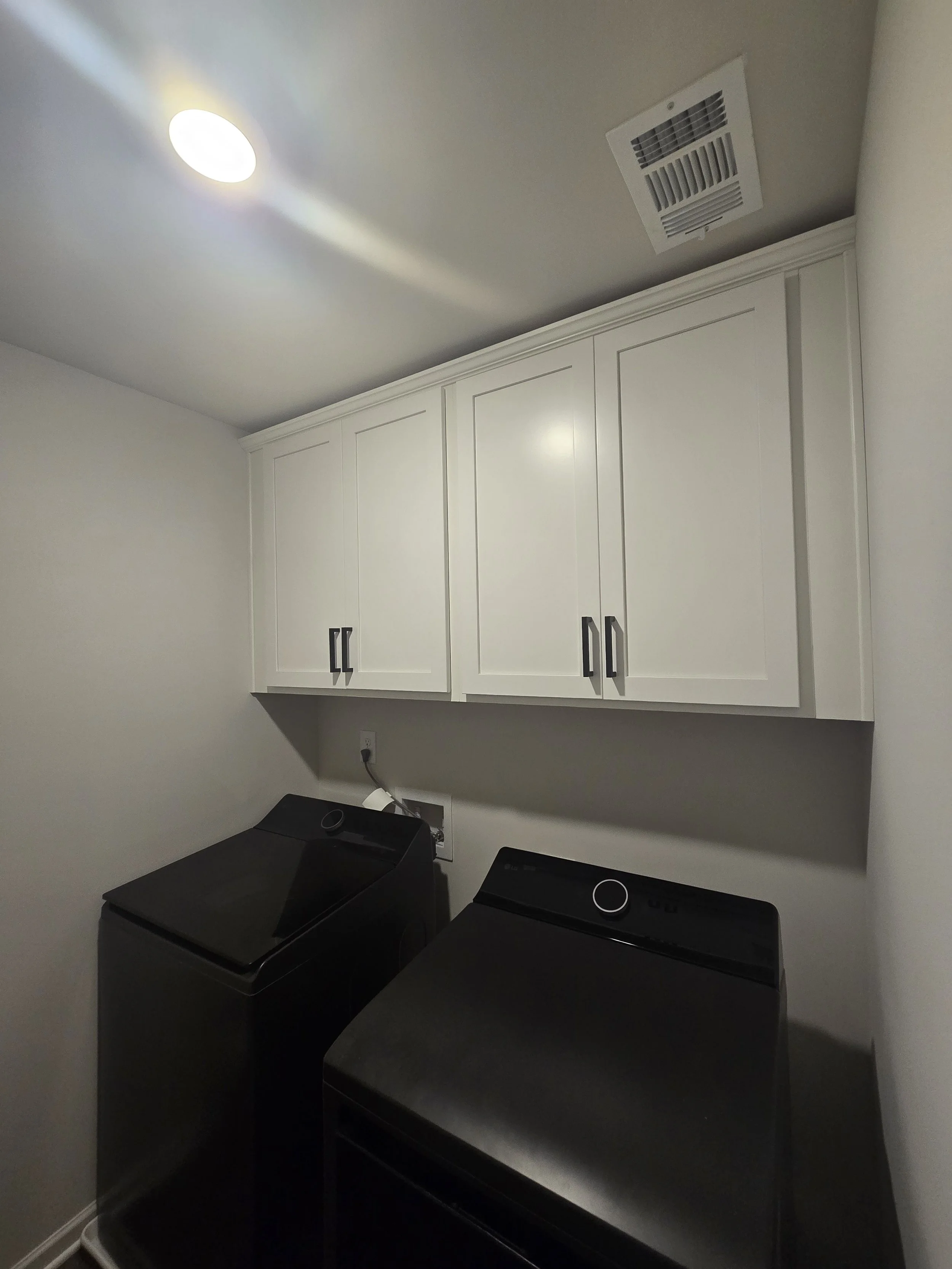 Laundry room with white cabinets above black washer and dryer, ceiling vent, ceiling light, and wall outlet.