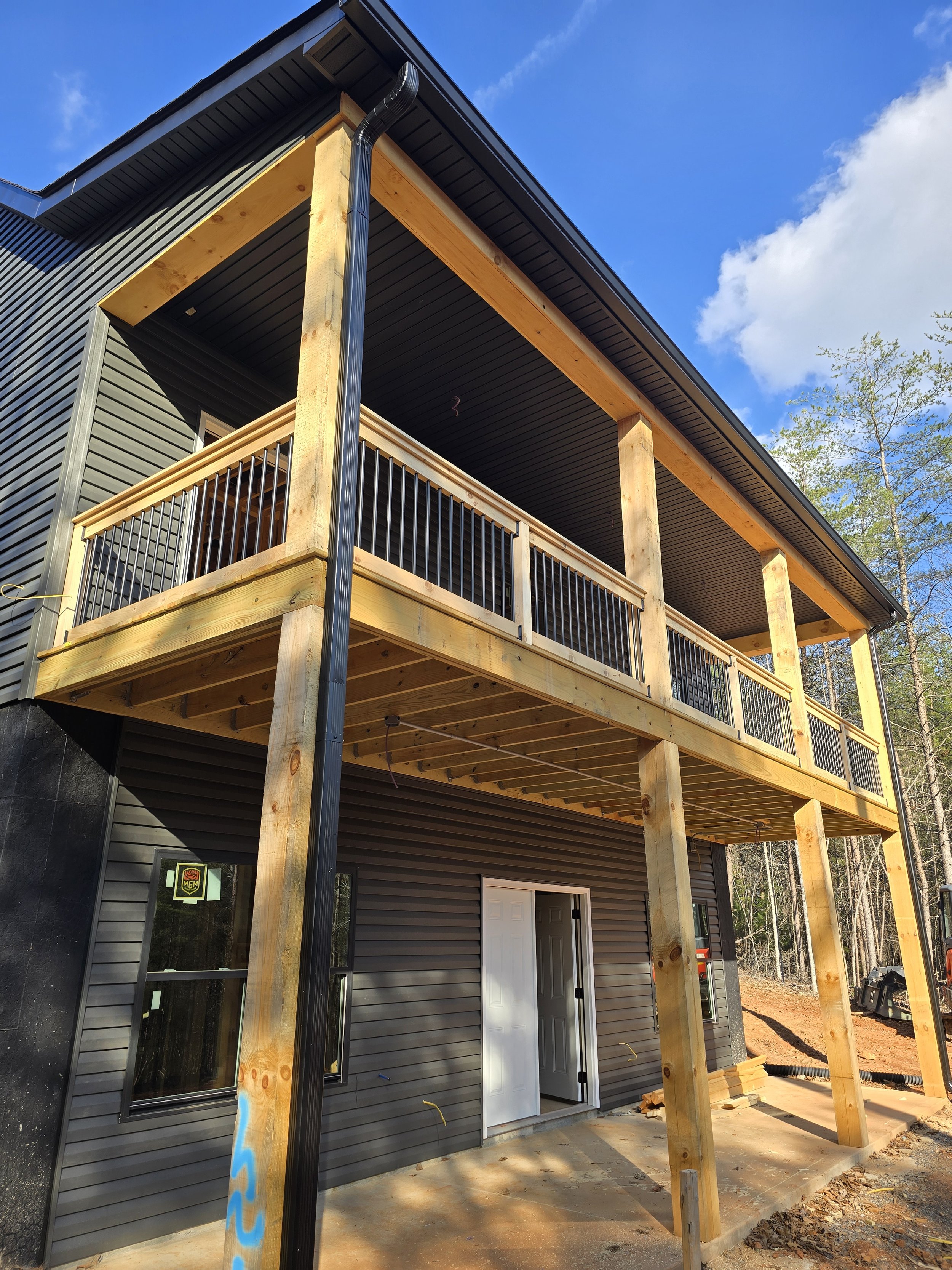 Exterior view of a house under construction with a new second story or porch being built with wooden beams and black metal railing, with a black metal downspout attached to the roof, large windows, and a partially open white door, set against a backg