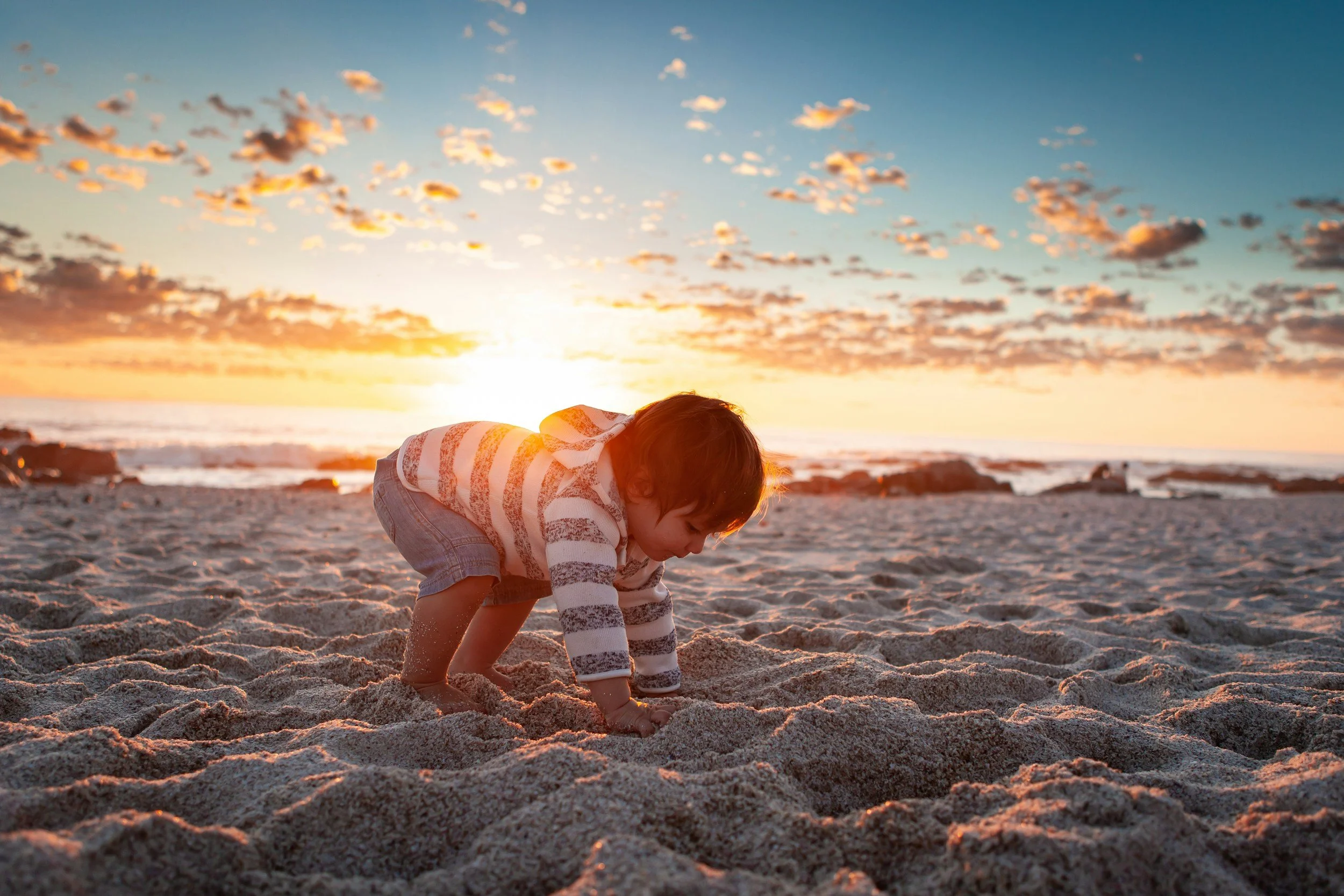A child crouching on sandy beach at sunset, with ocean and sky in background.