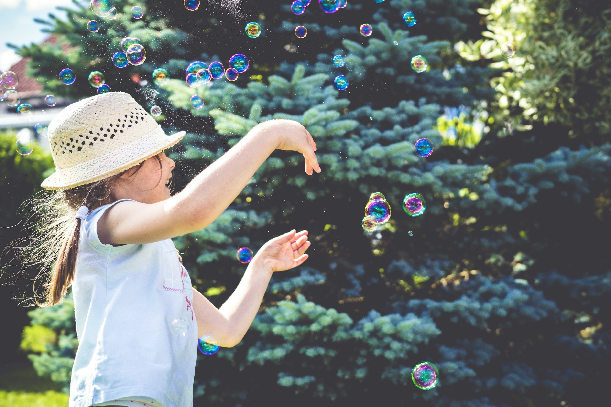 A young girl in a white sleeveless top and a straw hat playing with soap bubbles outdoors, with a bushy evergreen tree in the background.