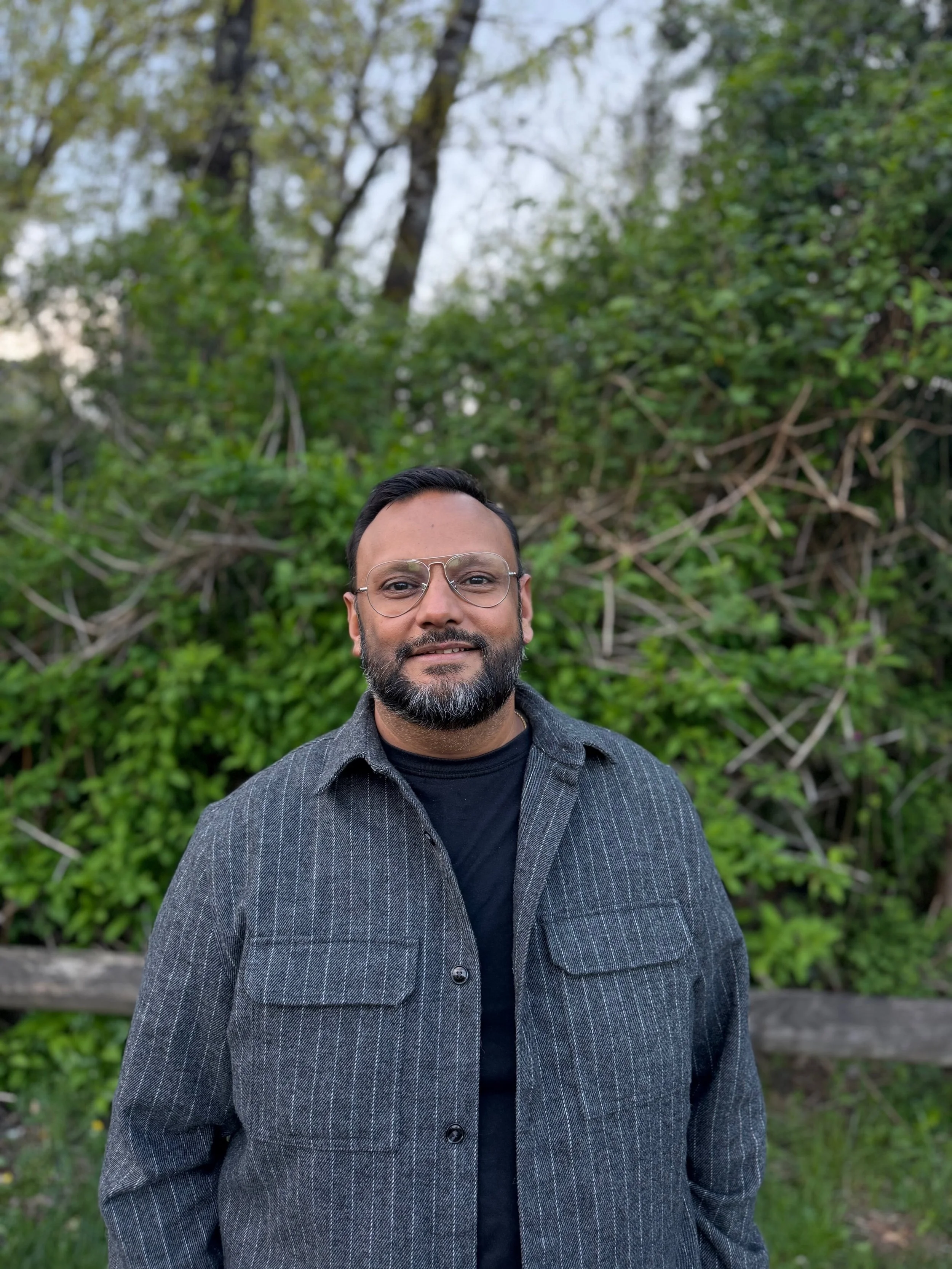 A man with glasses, a beard, and a mustache standing outdoors in front of green trees and bushes, wearing a dark gray pinstripe coat over a black shirt.