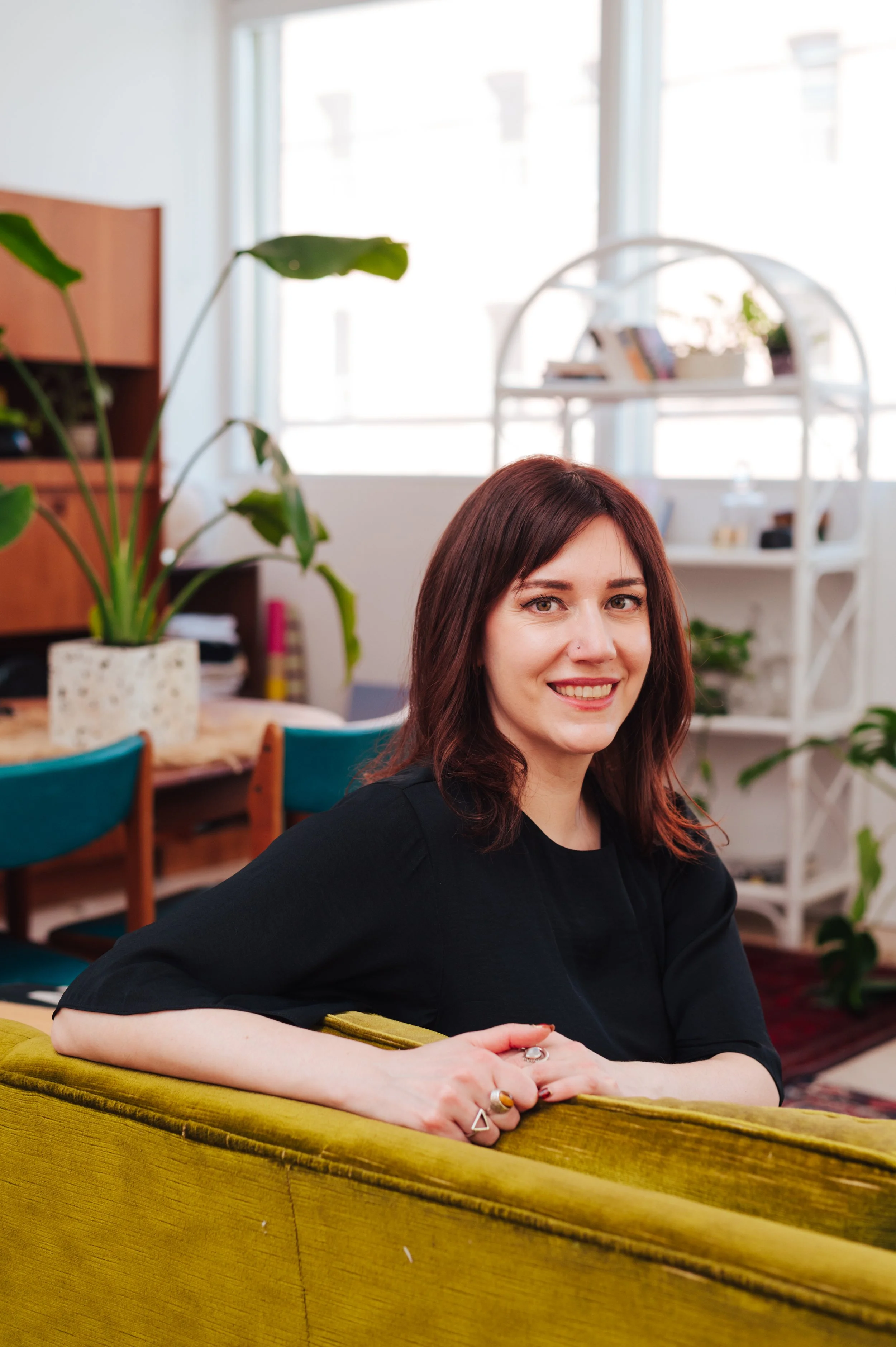 A woman (Dr Amy Reichelt) with shoulder-length dark red hair, wearing a black top, sitting on a mustard-colored couch in a bright, cozy room with plants and white furniture.