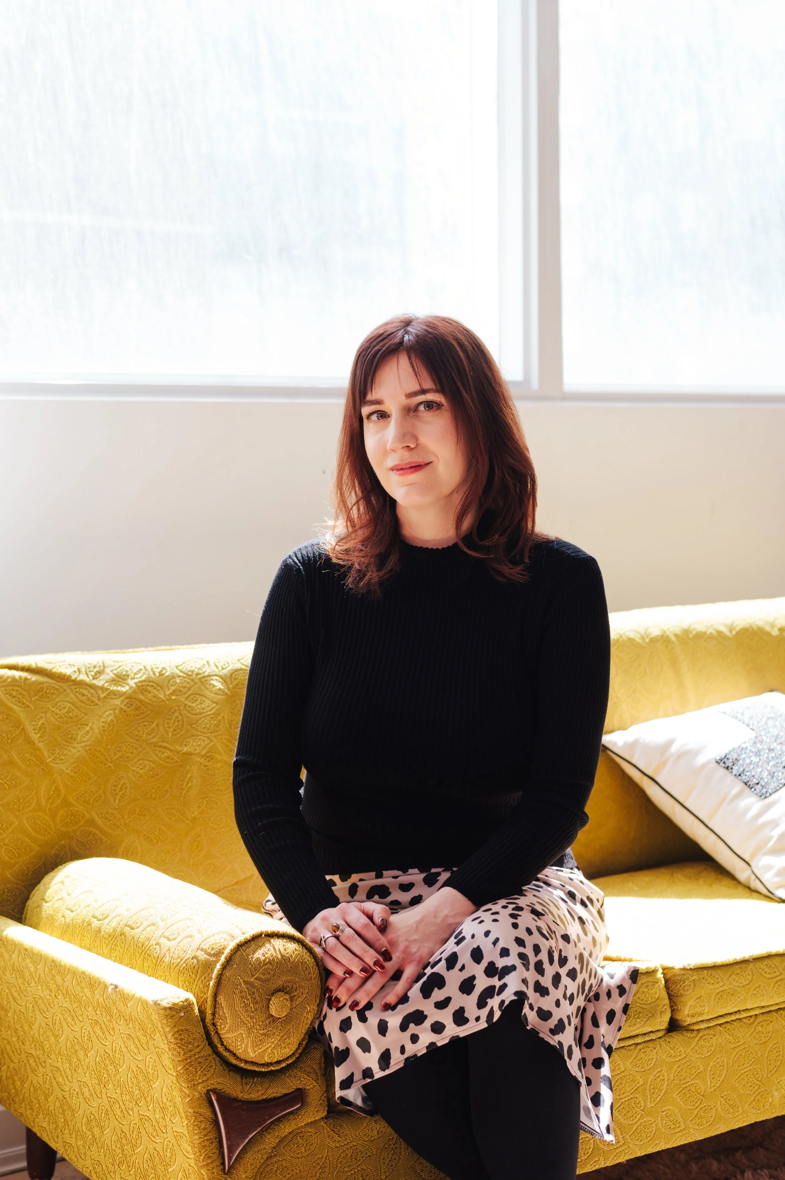 Woman (Dr Amy Reichelt) with brown hair in a black top and animal print skirt sitting on a yellow patterned couch near a window.