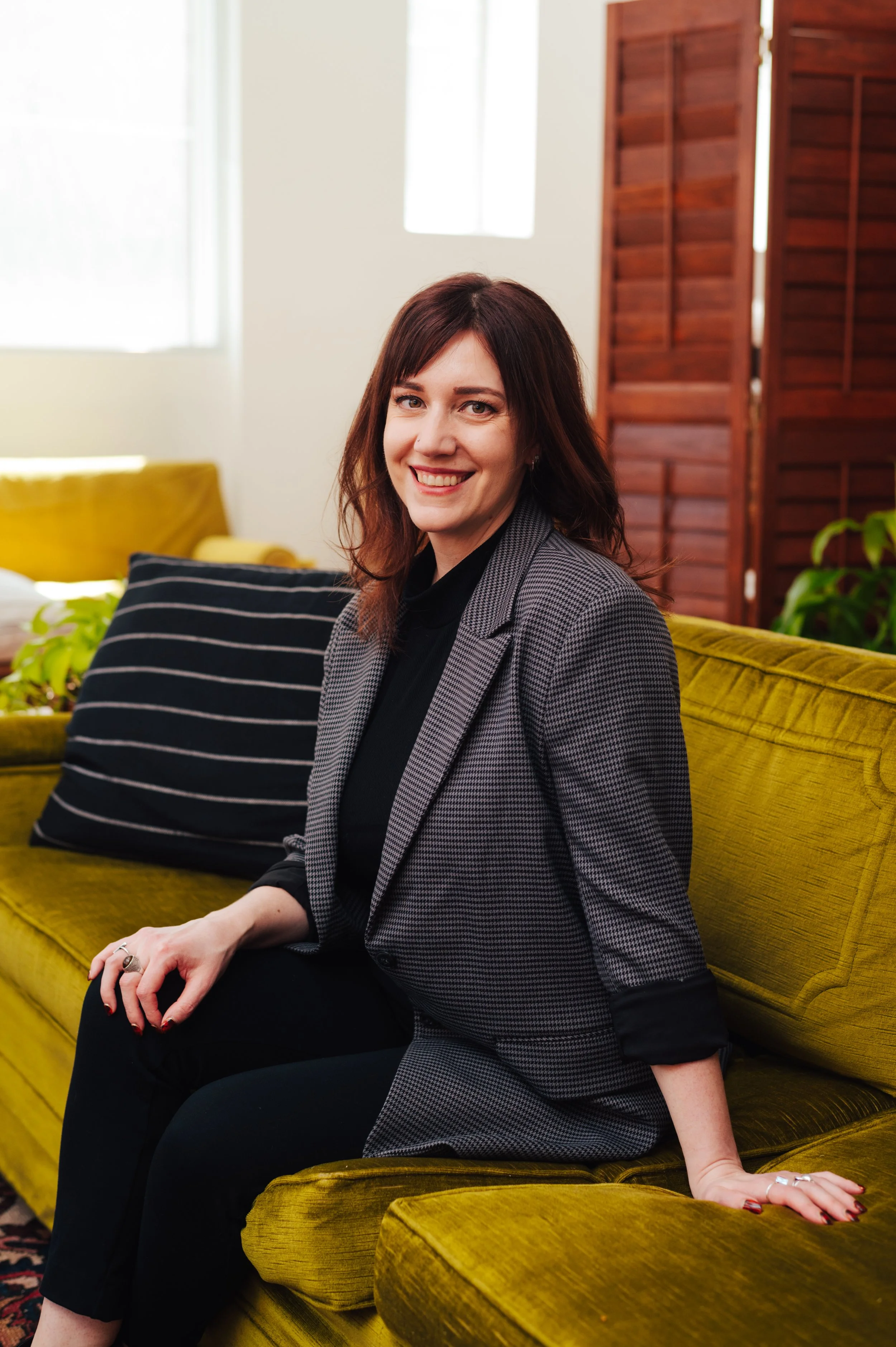 A woman (Dr Amy Reichelt) with shoulder-length brown hair, wearing a black shirt and gray checkered blazer, sitting on a yellow velvet sofa, smiling at the camera, in a room with wooden window blinds and green plants.