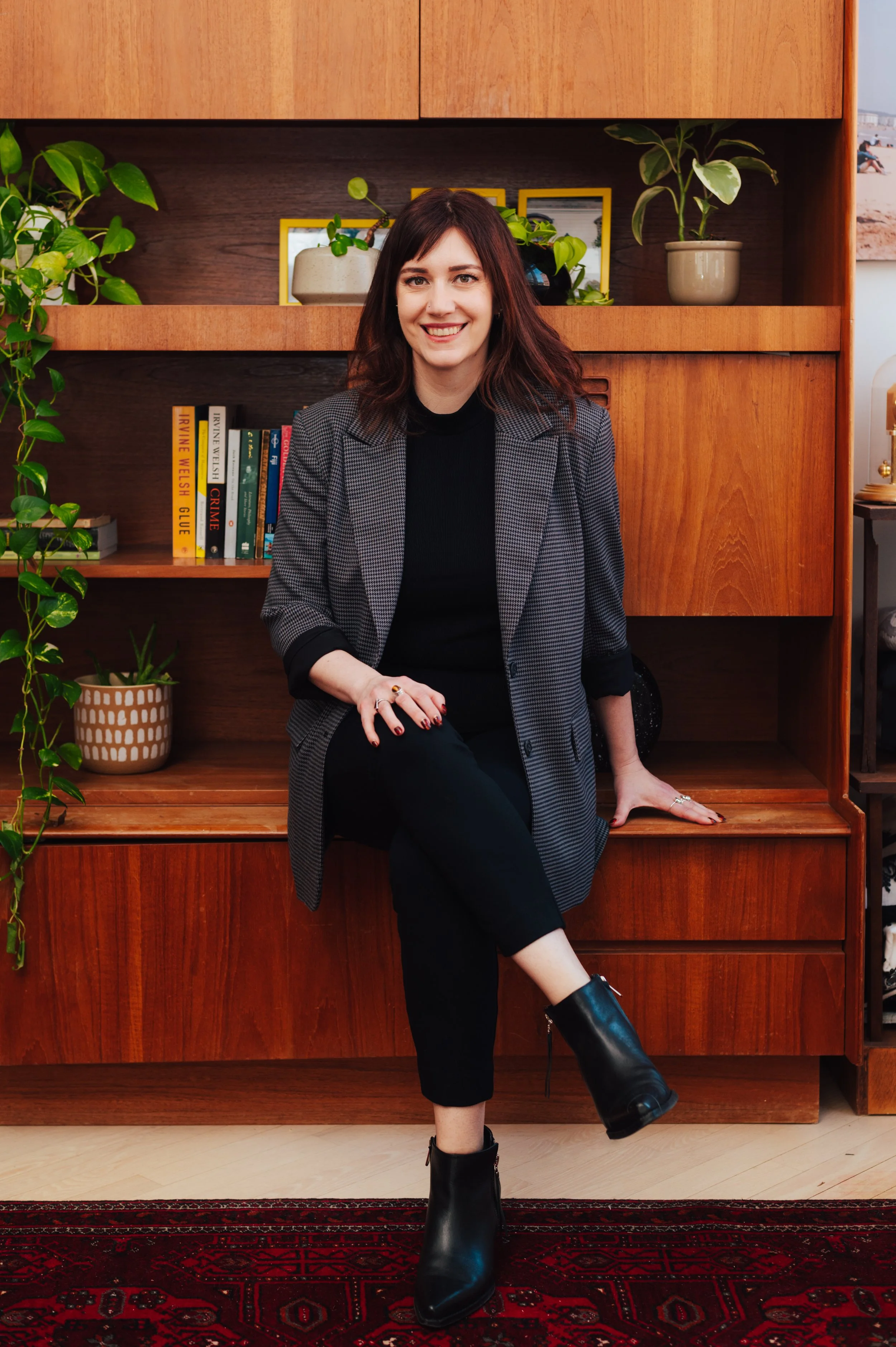 Woman (Dr Amy Reichelt) sitting in front of a wooden bookshelf with books and potted plants, smiling.