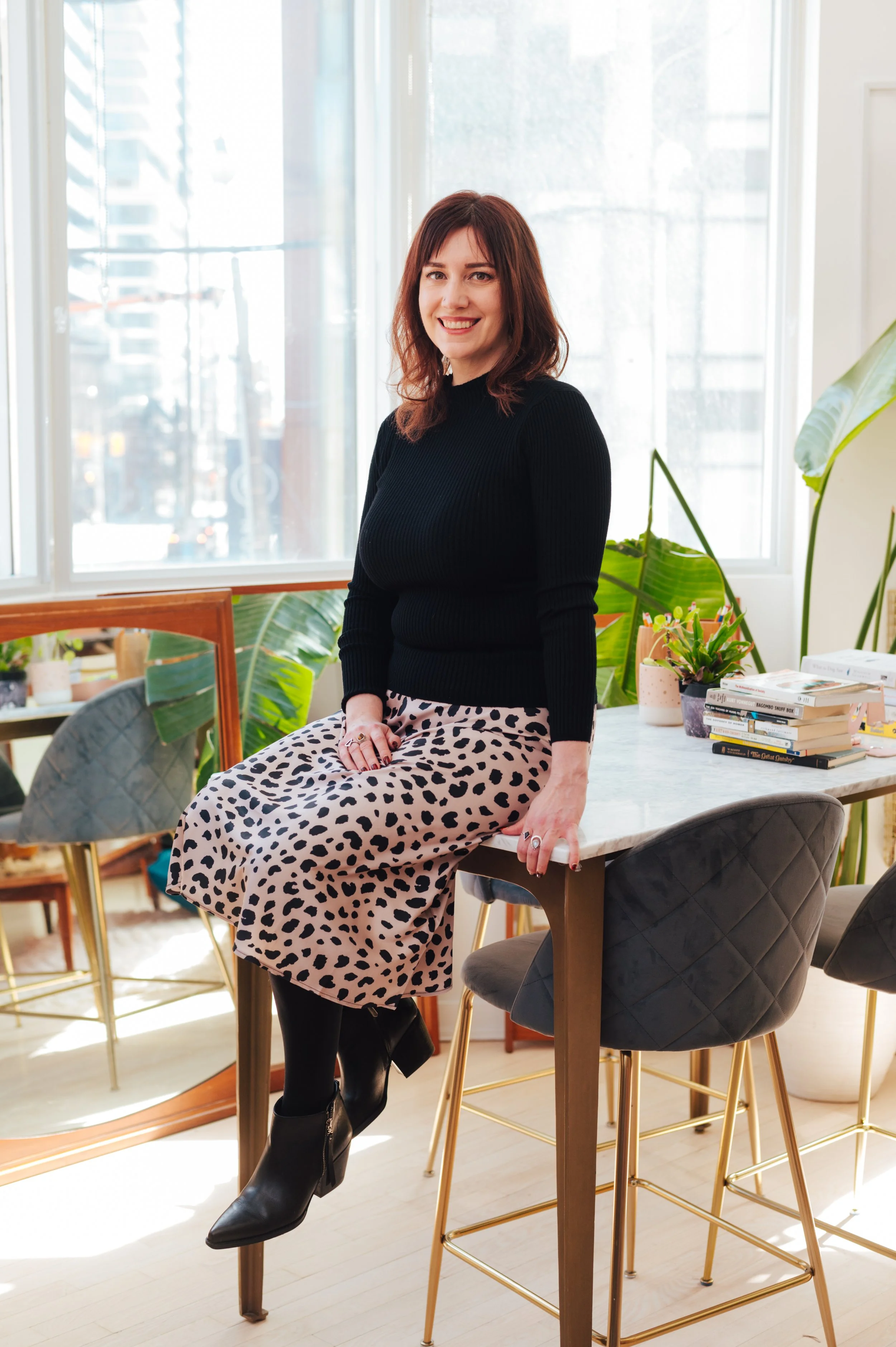 A woman with shoulder-length brown hair in a black top and animal print skirt, sitting on the edge of a dining table in a bright room with large windows and indoor plants.